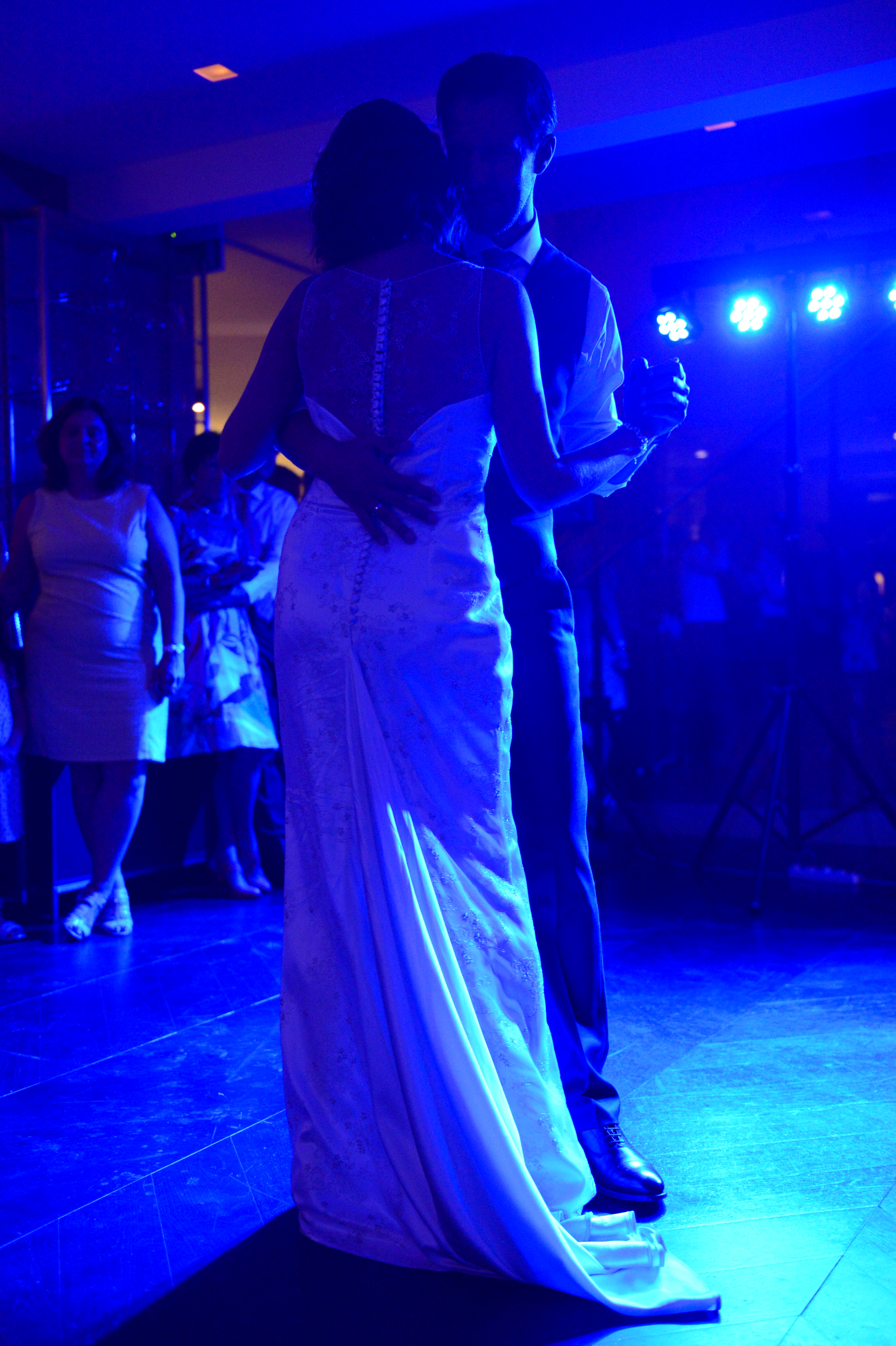 The bride and groom share their first dance, embracing closely on the dance floor under blue lighting.