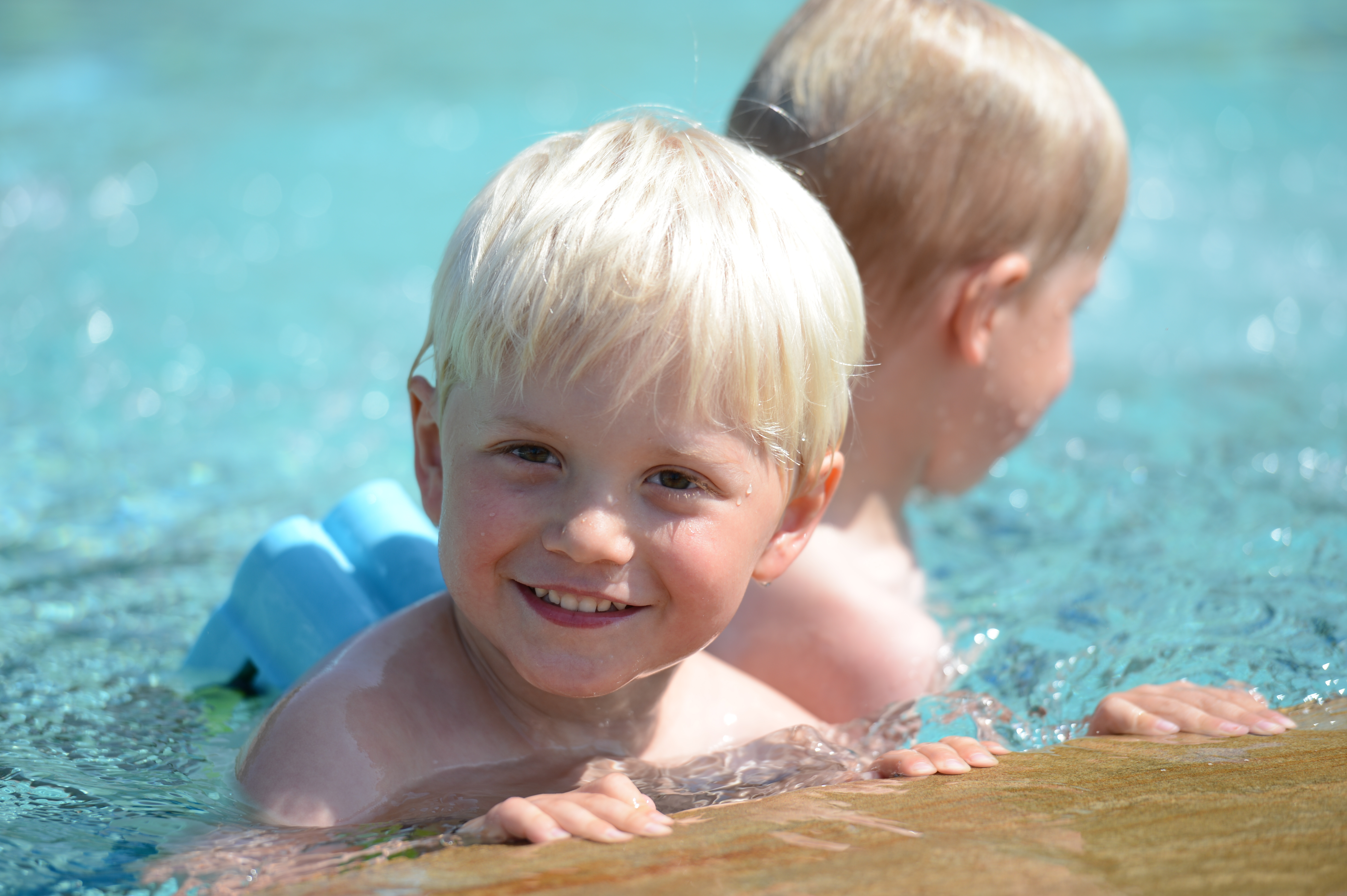 Two young children swimming in a pool, one smiling at the camera while the other faces away.