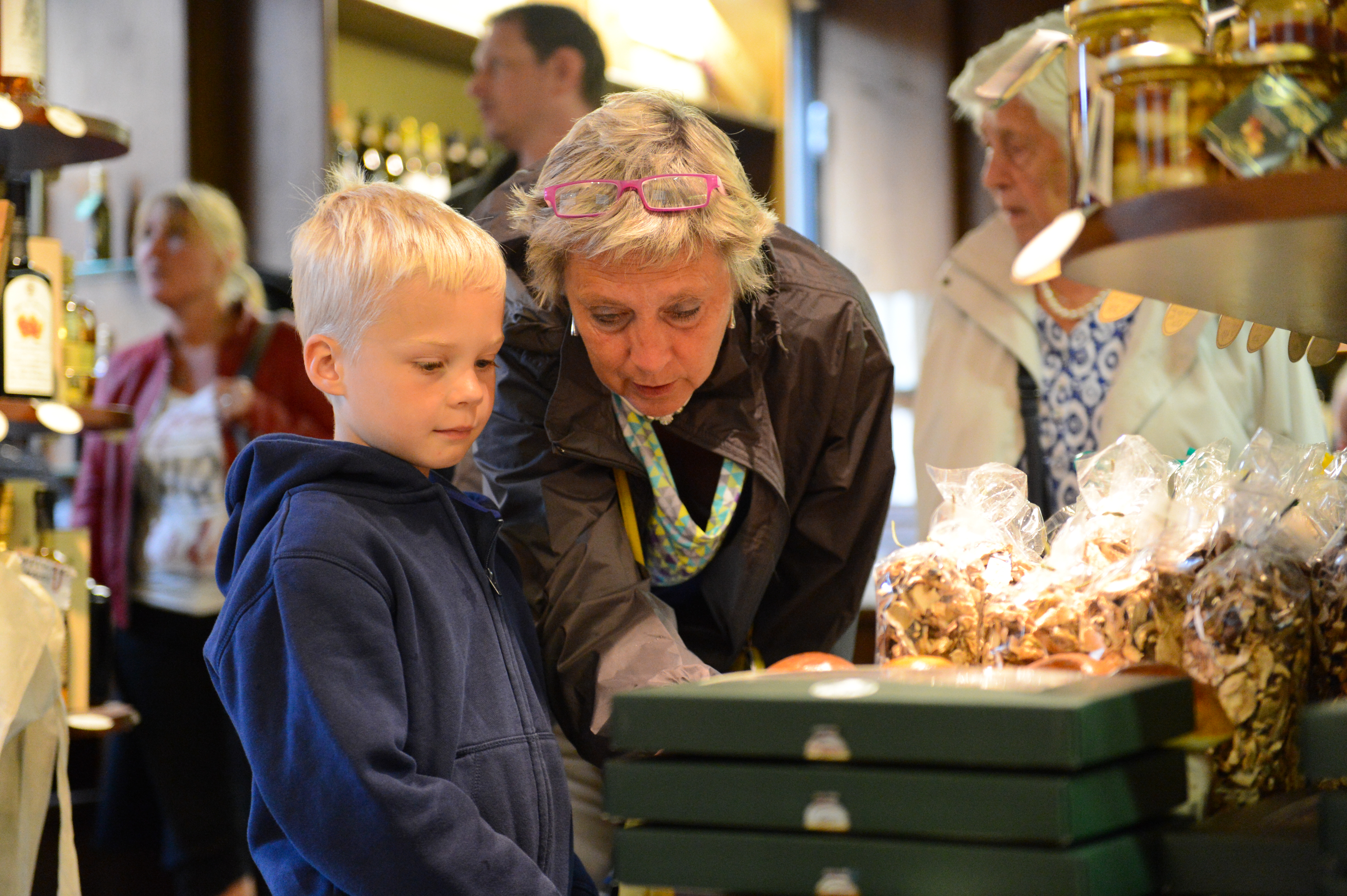 An older woman and a young boy look at packaged goods in a store, with other shoppers in the background.