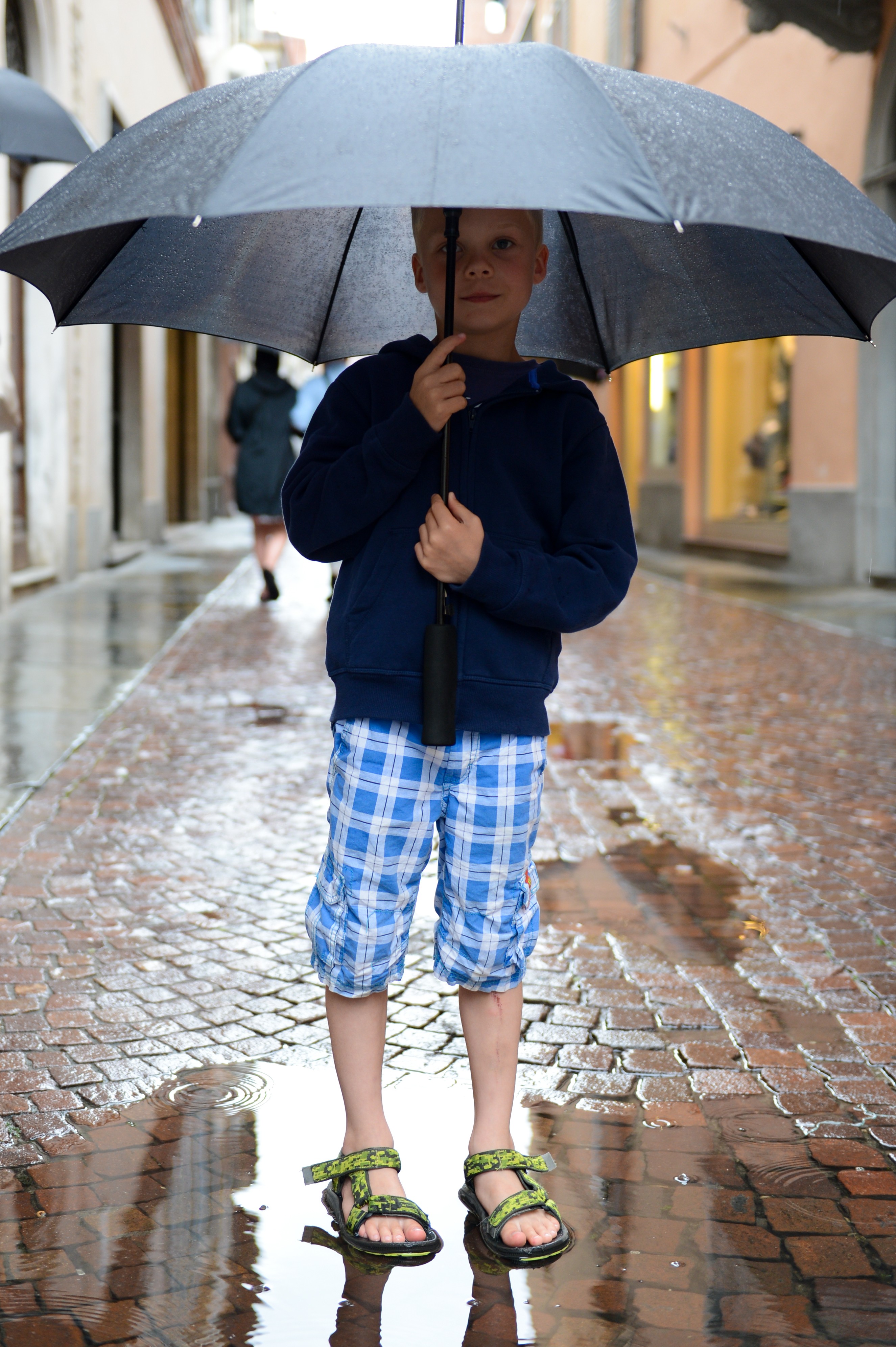 A young boy stands on a wet cobblestone street, holding a large umbrella in the rain.