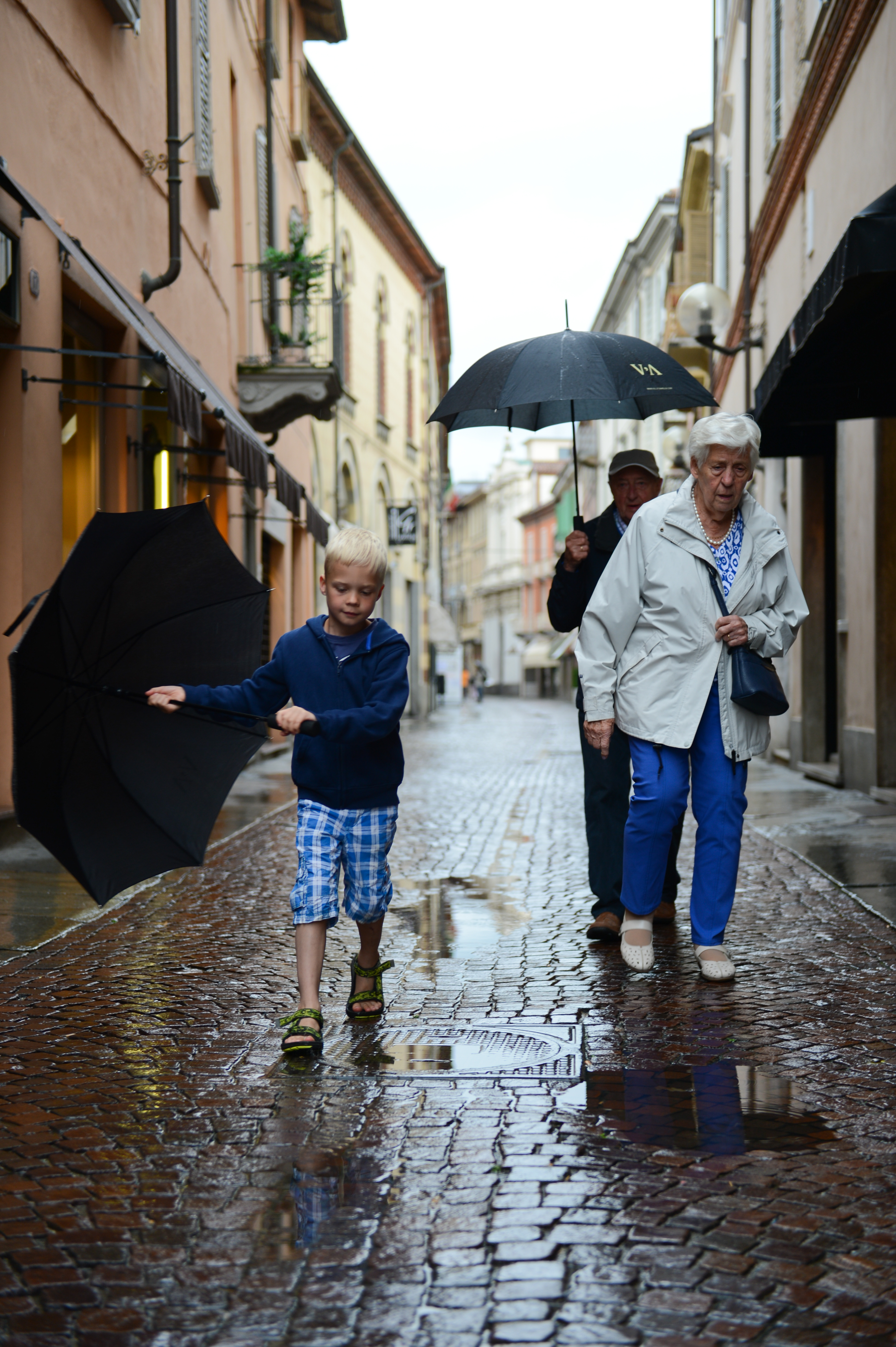 A young boy and two older adults walk on a wet cobblestone street, holding umbrellas on a rainy day.