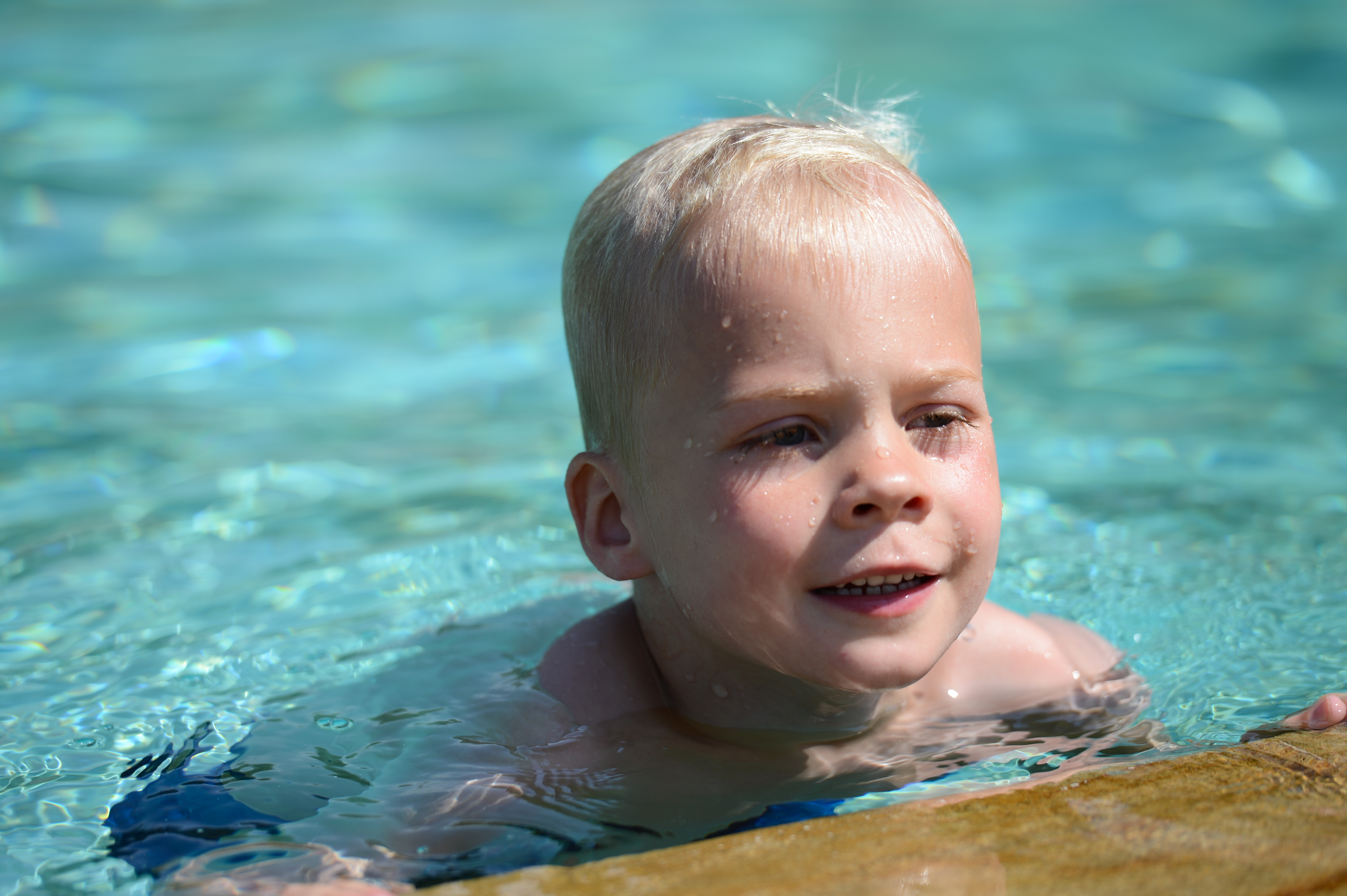 A young child with wet hair swims in a pool, holding onto the edge and looking forward.