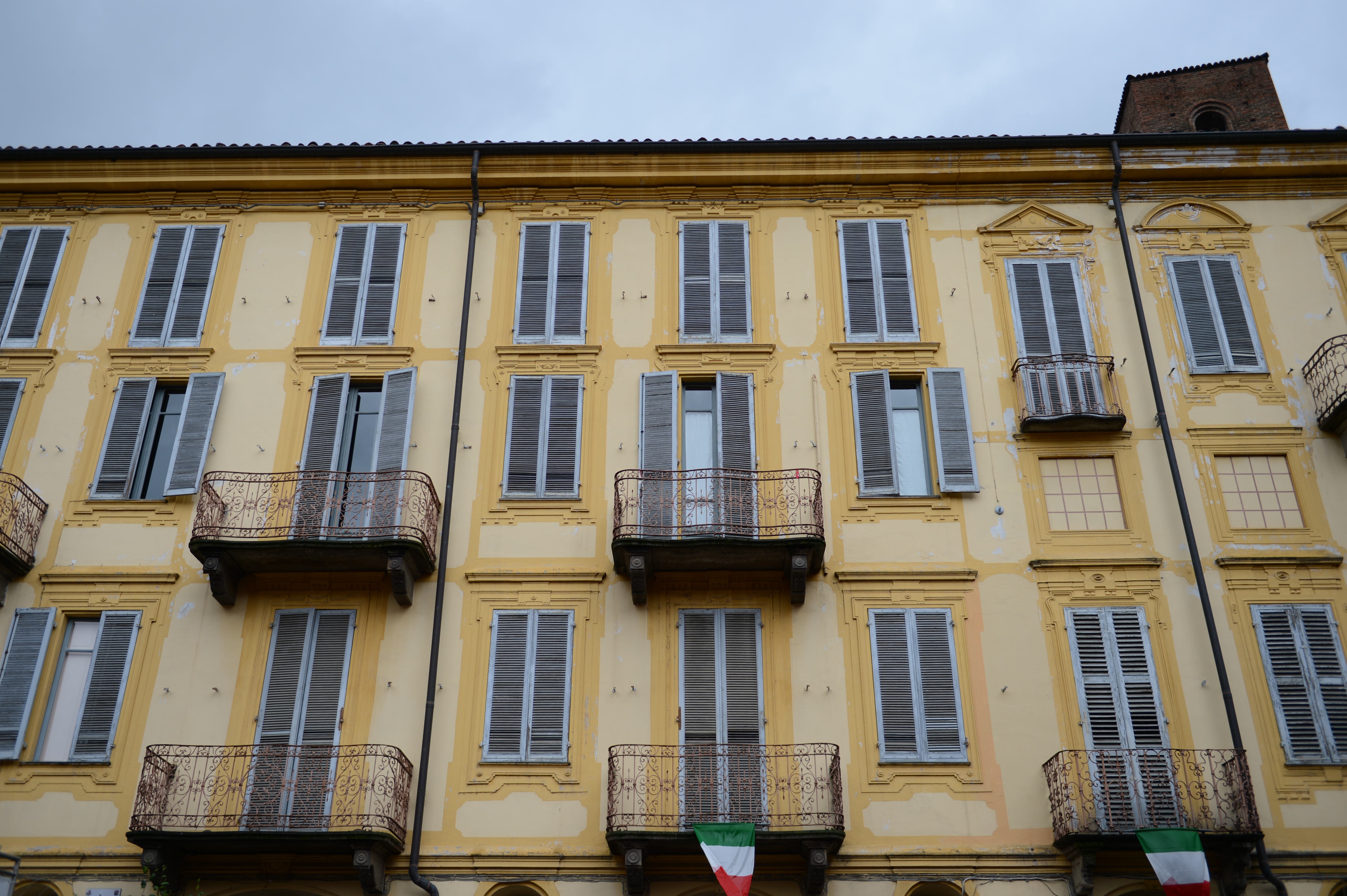 A yellow building with multiple windows and balconies, some with open shutters, and Italian flags hanging below.