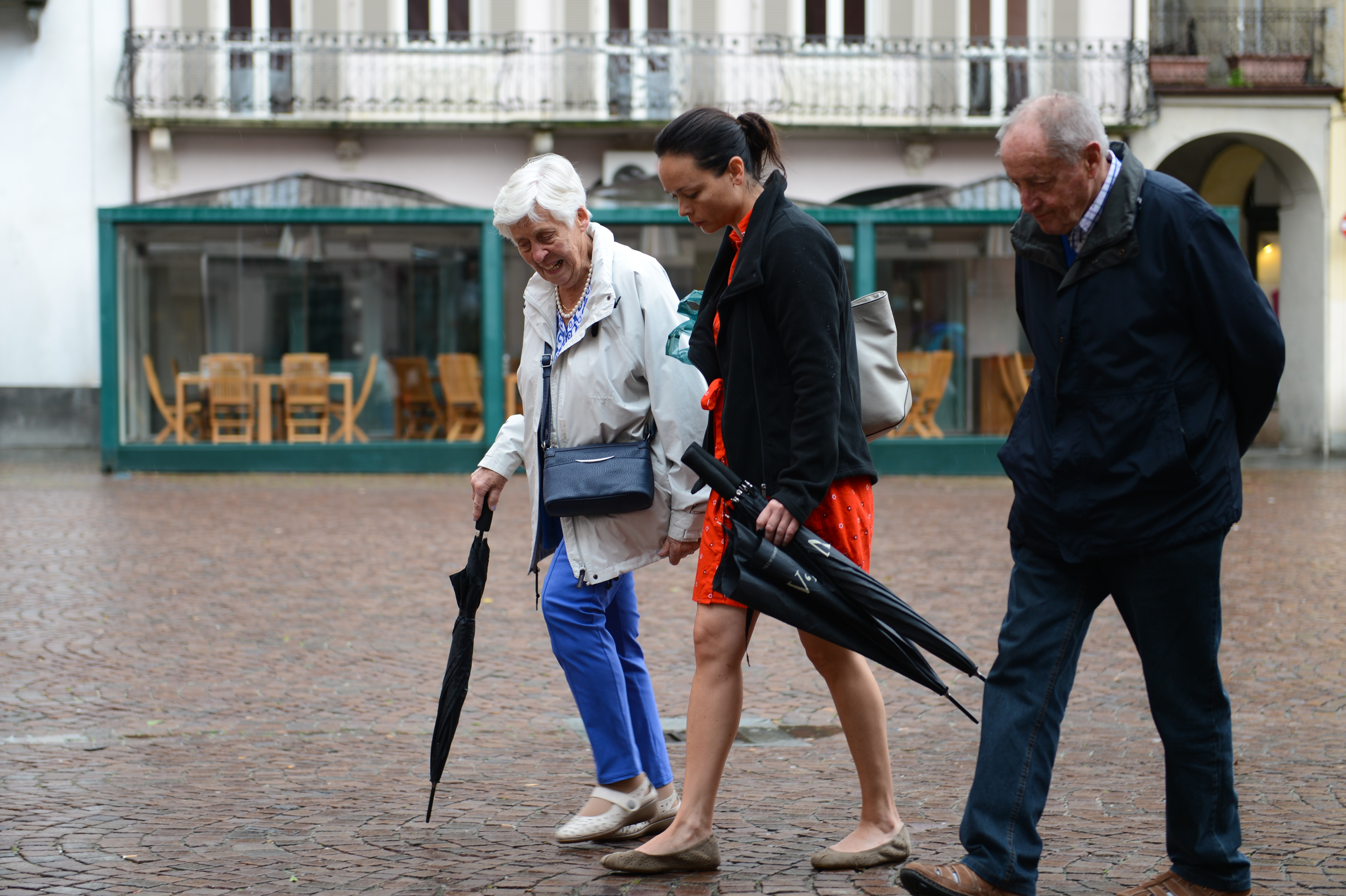 Three people walk on a wet cobblestone street, carrying umbrellas and engaged in conversation.