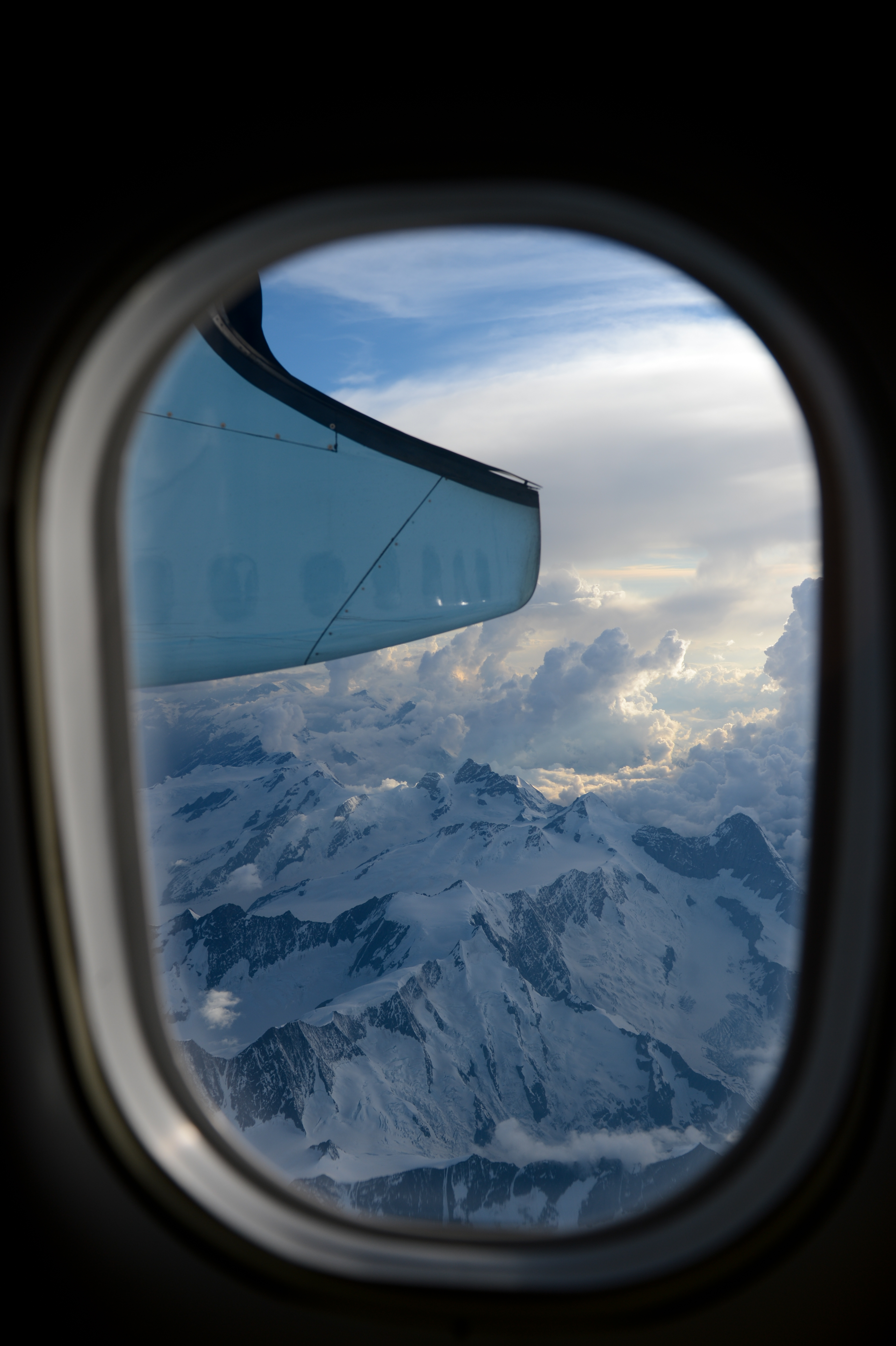 View from an airplane window showing snow-covered mountains and clouds, with part of the wing visible.