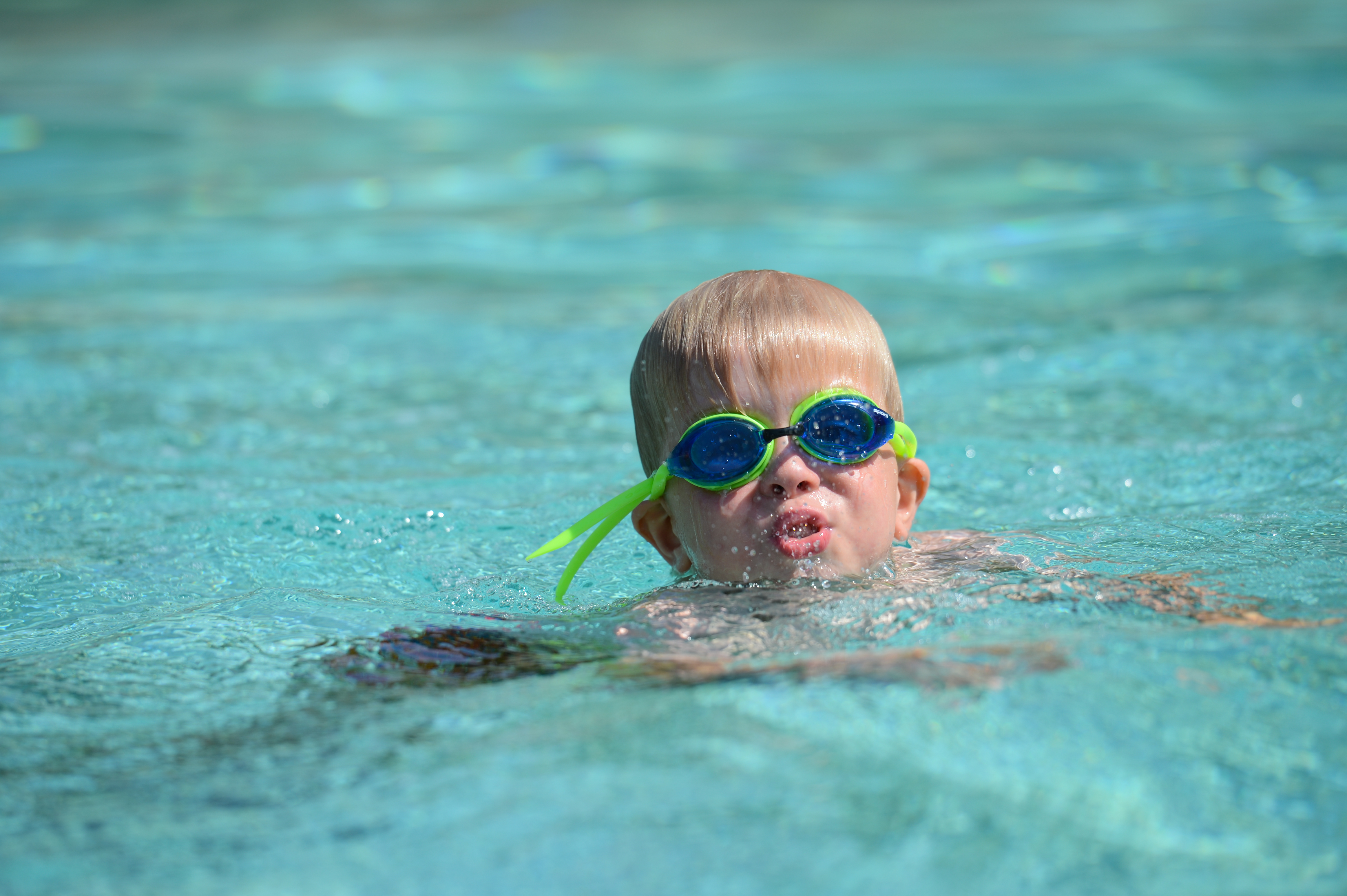 A young child wearing goggles swims in a pool, with their face partially above the water.