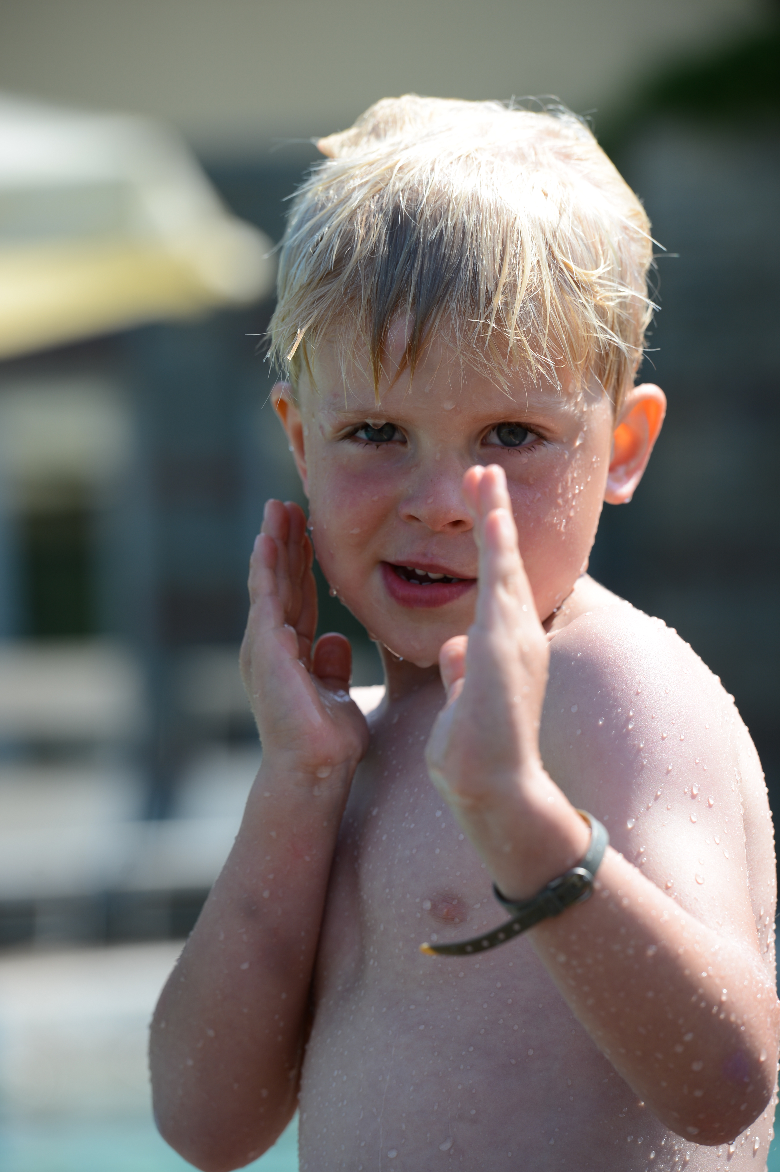 A young child with wet hair and skin stands near water, raising their hands playfully.