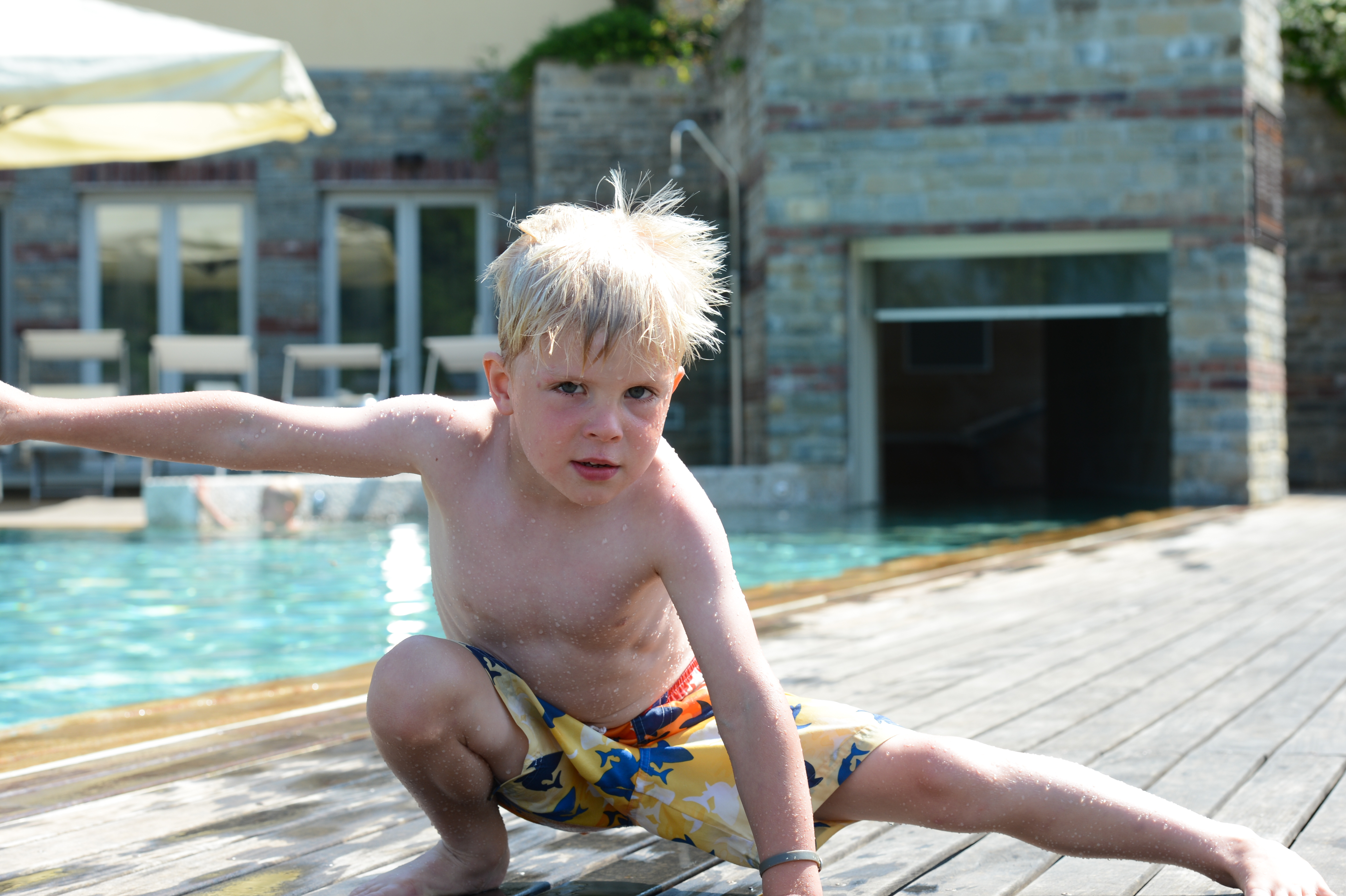 A young boy in swim trunks crouches playfully on a wooden deck near a pool, extending his arm.