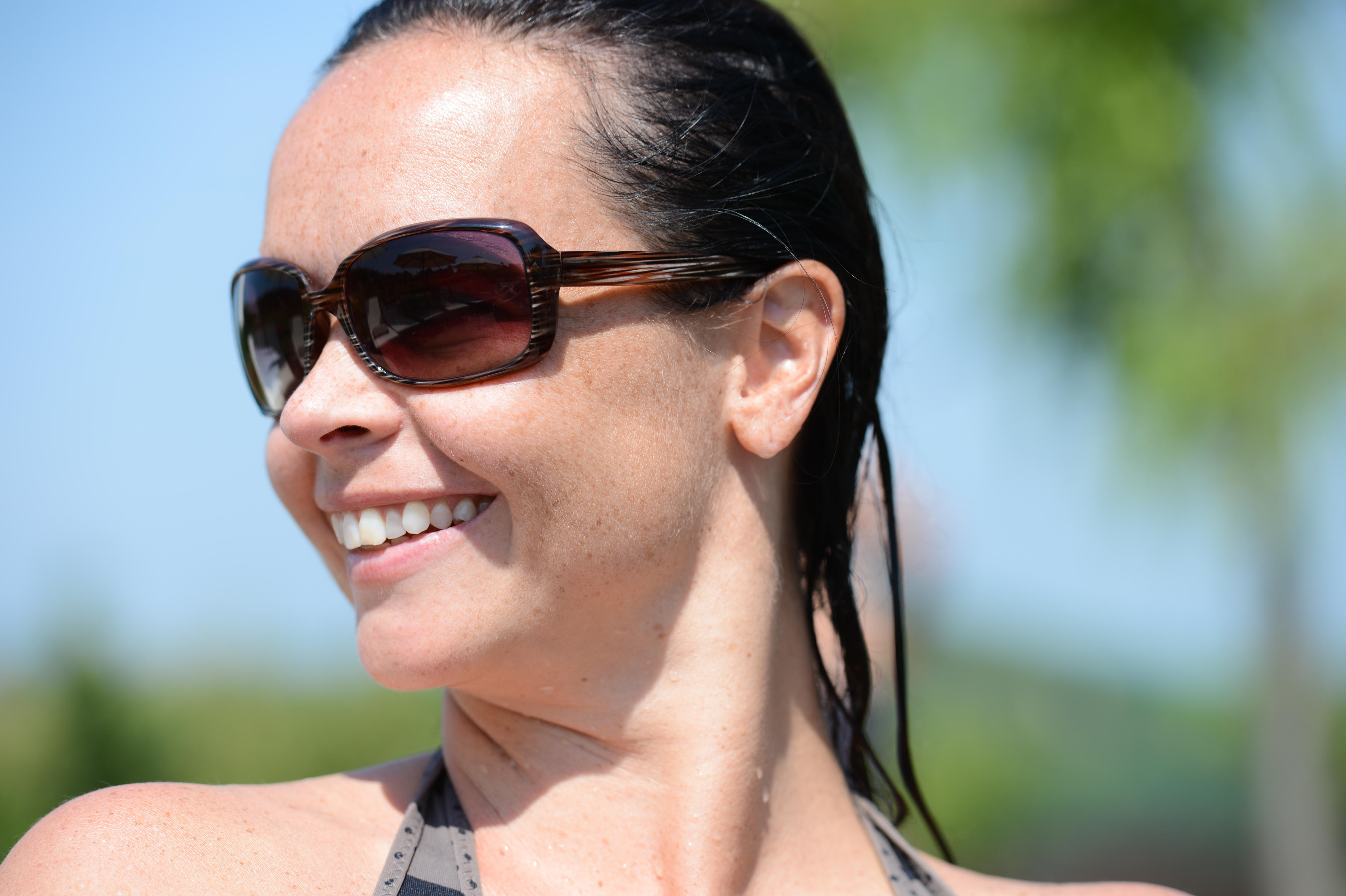 A woman with wet hair and sunglasses smiles while looking to the side in an outdoor setting.