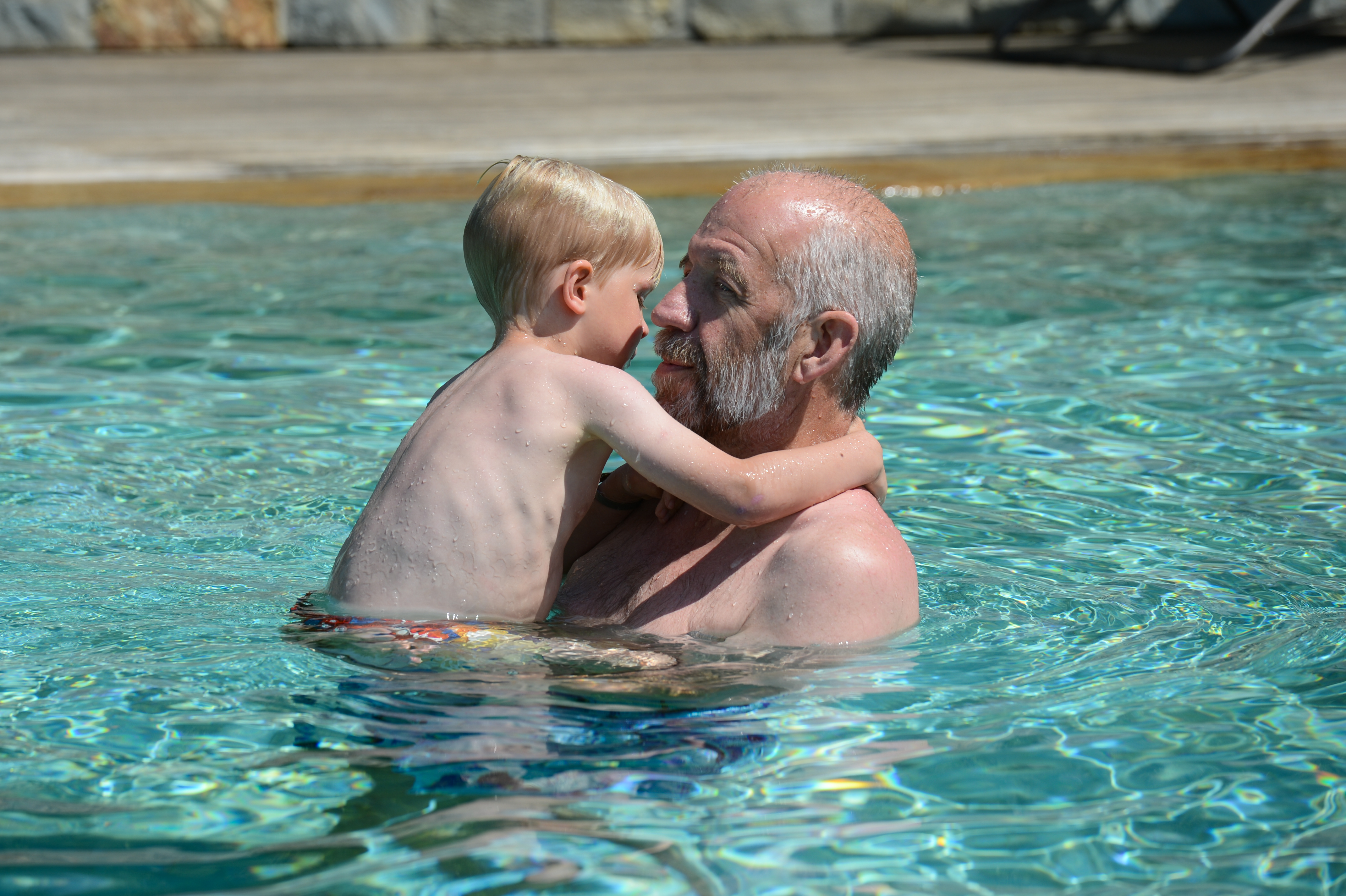 An older man holds a young child in a swimming pool, both looking at each other closely.