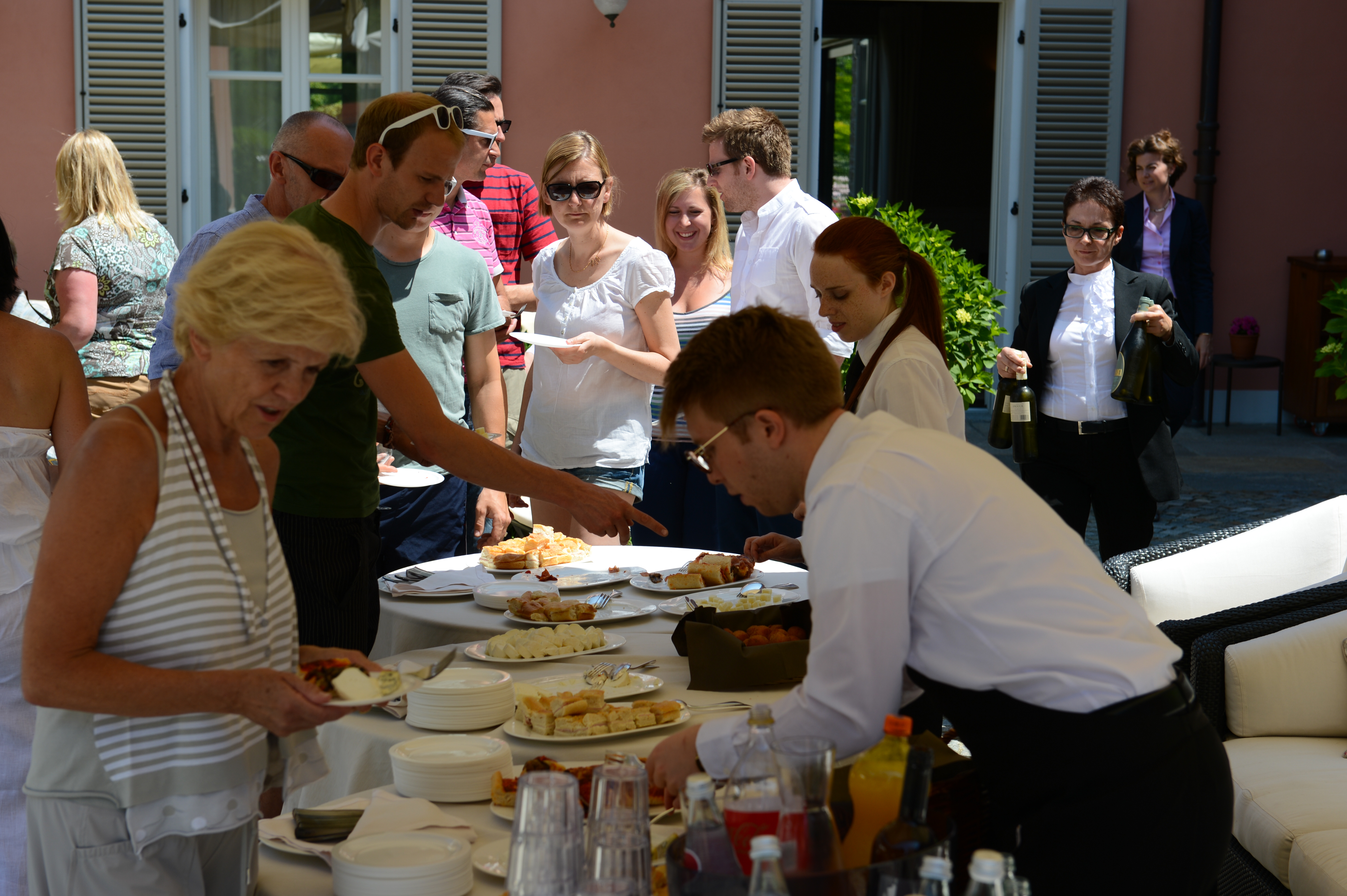 Guests gather around a buffet table at an outdoor wedding reception, selecting food while servers assist.