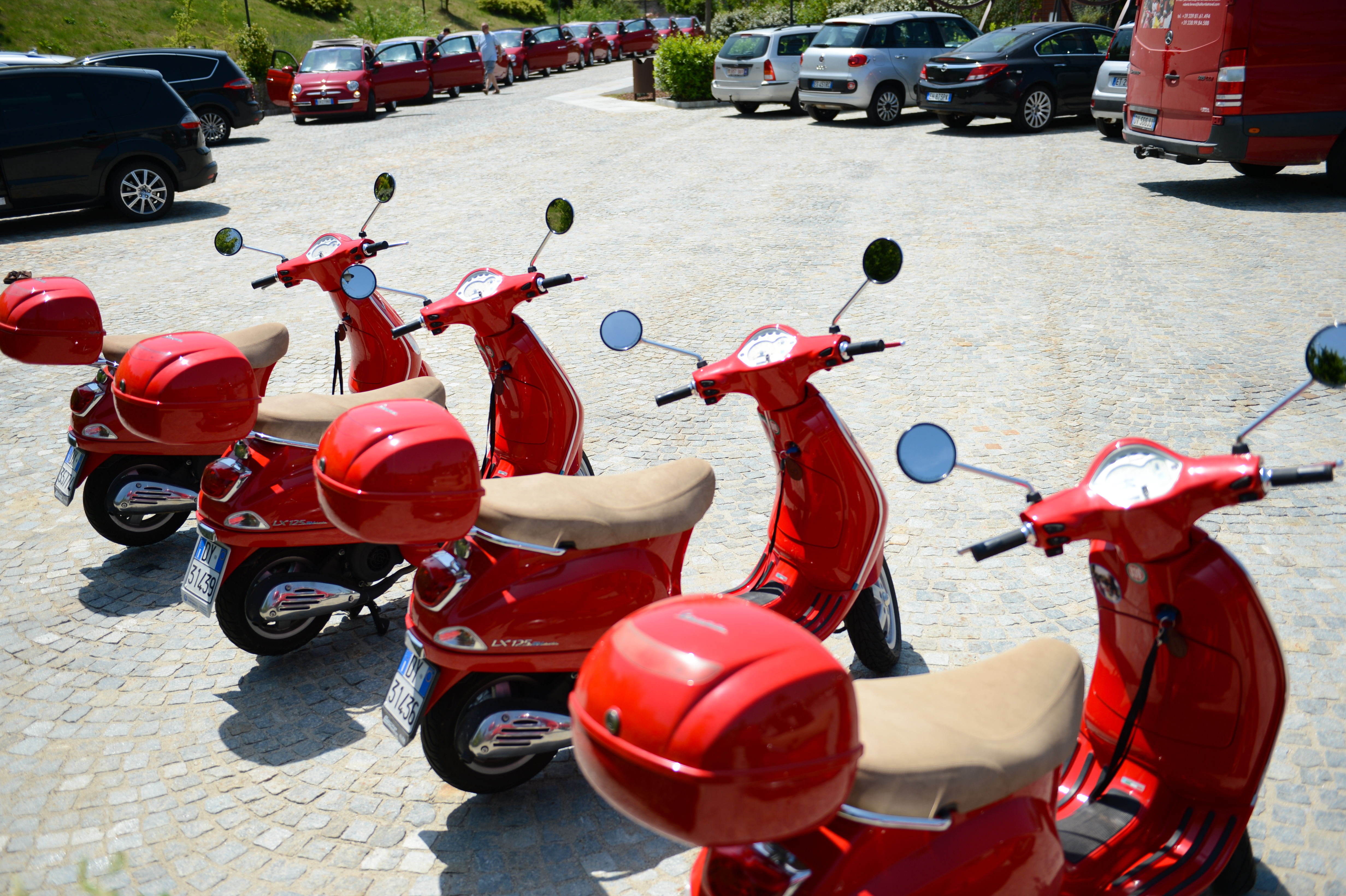 Five red scooters with beige seats are parked in a row on a cobblestone surface, with cars in the background.
