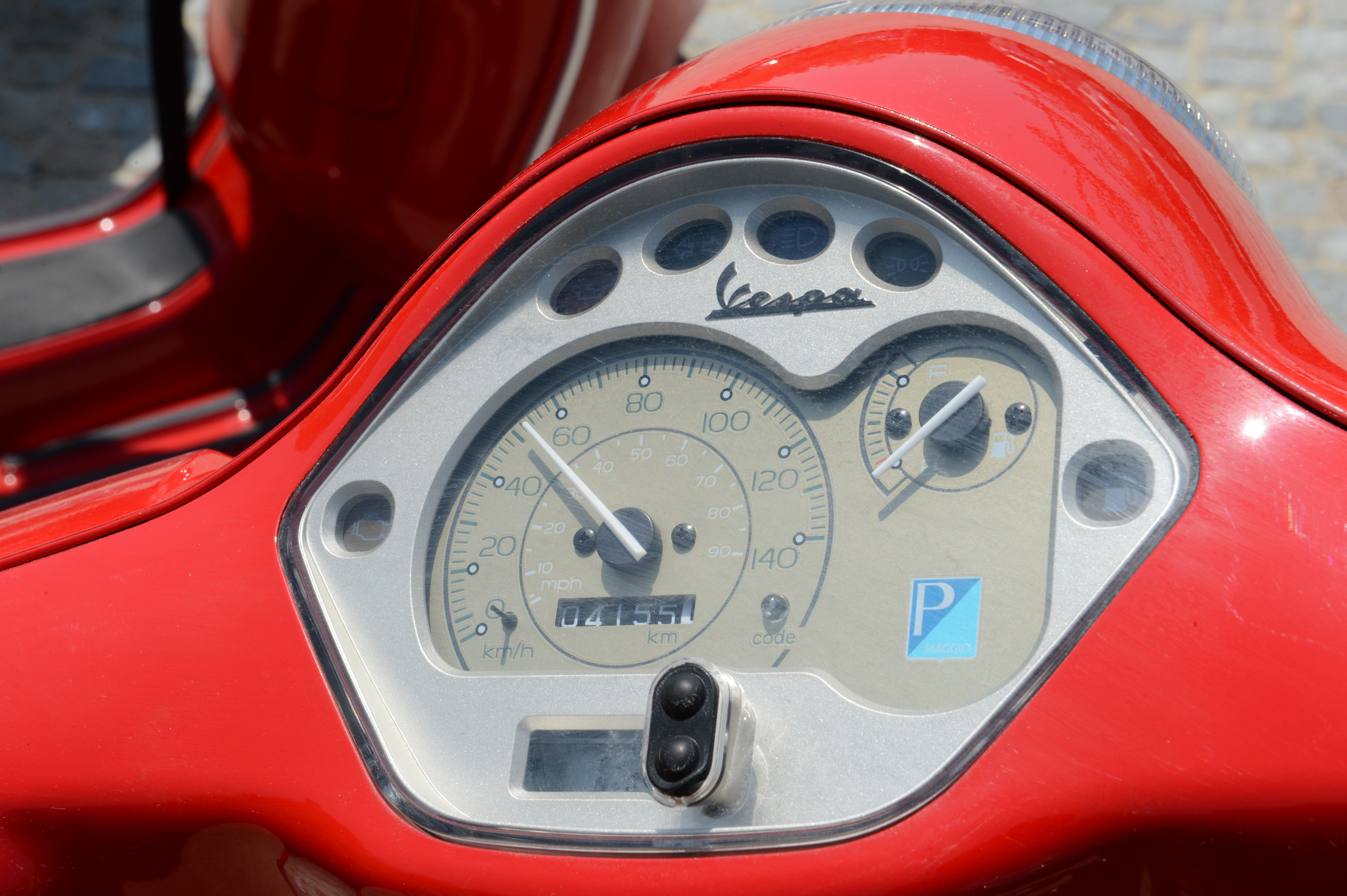 Close-up of a red Vespa scooter's speedometer and fuel gauge, showing the odometer reading and parking sticker.