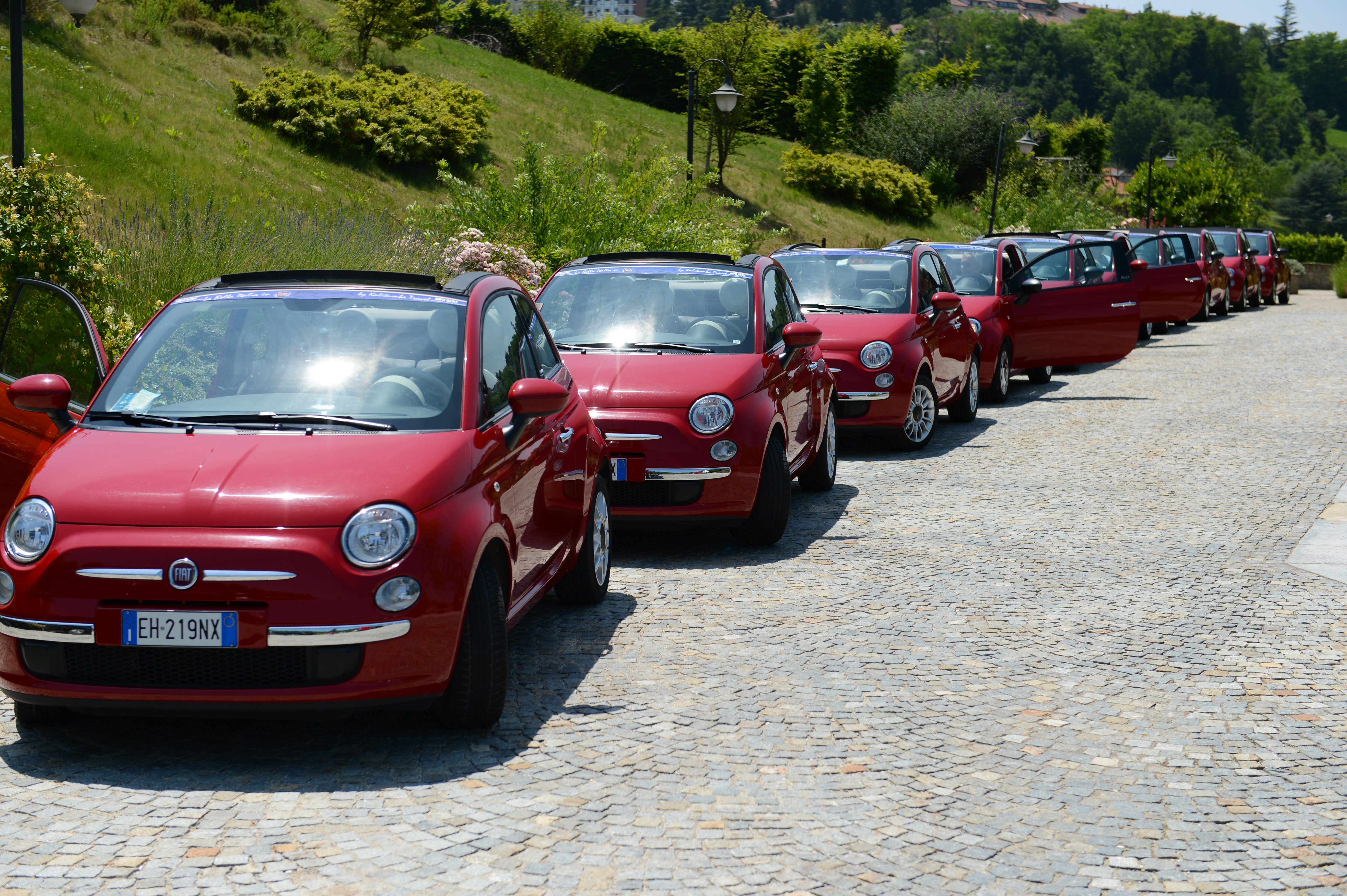 A row of red Fiat 500 cars with open doors, parked on a cobblestone path at a wedding event.