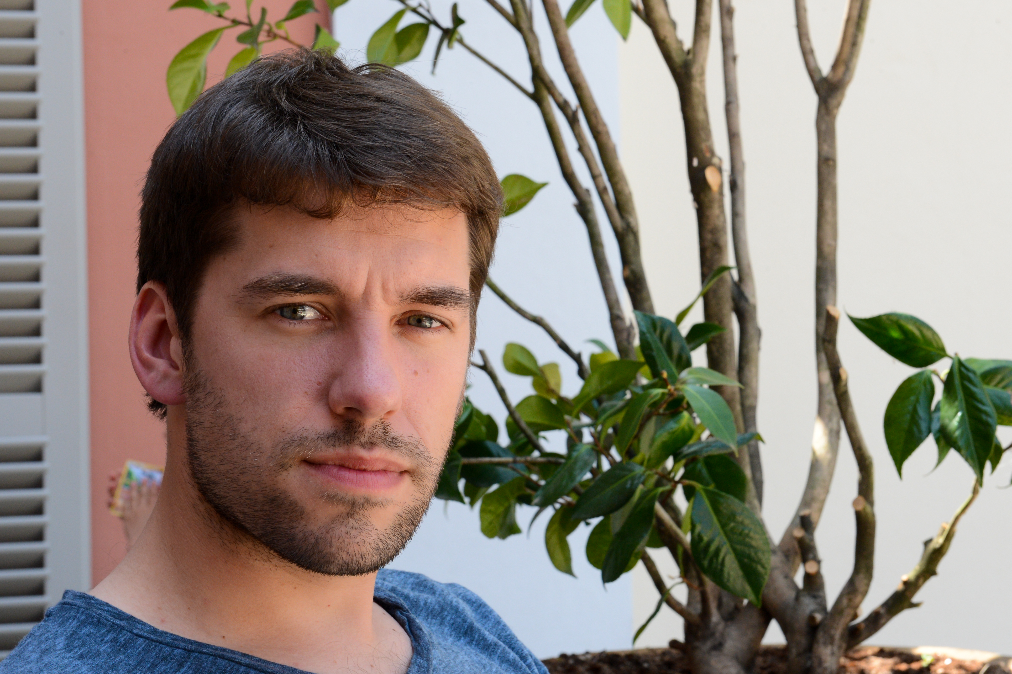 A man with short brown hair and a beard looks at the camera while sitting outdoors near a plant.