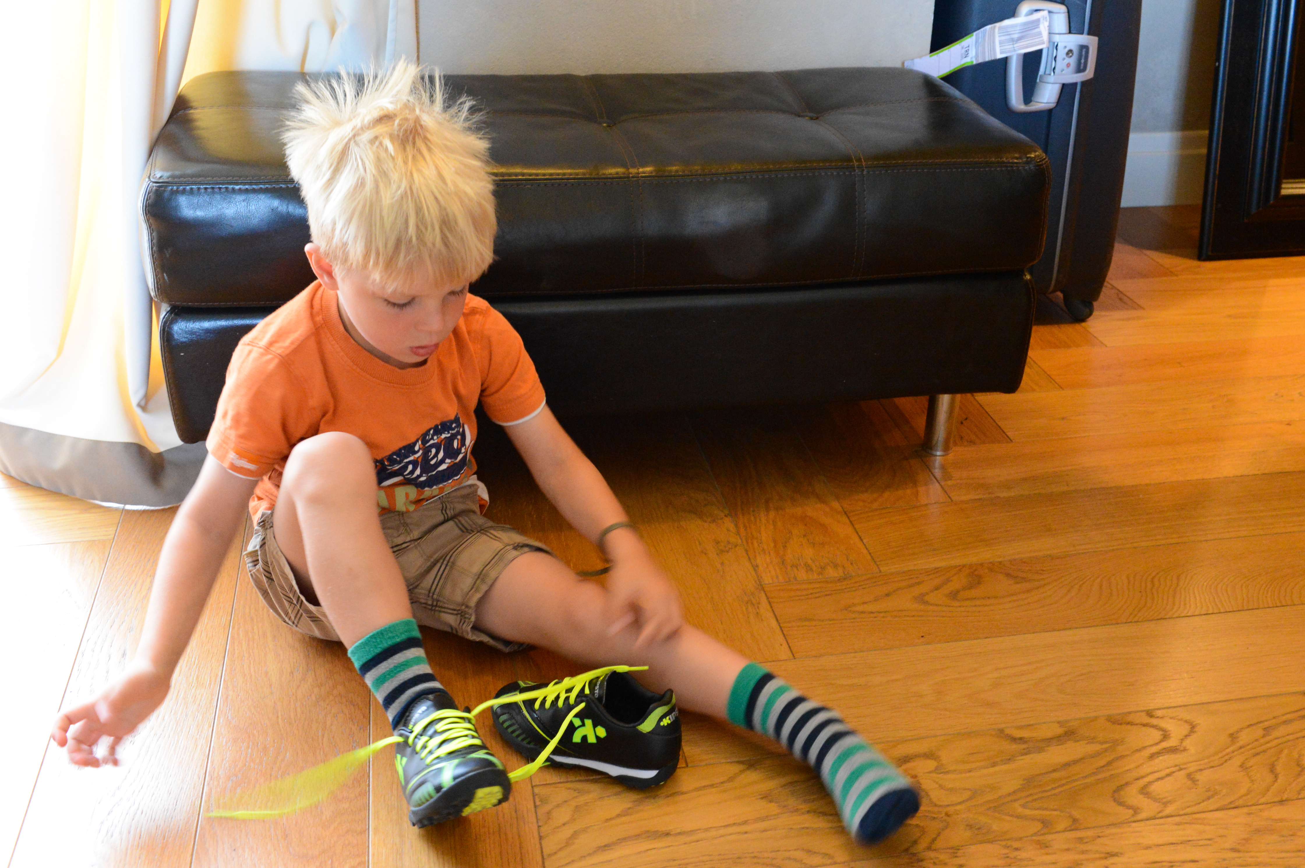 A young boy sits on the floor, tying the laces of his black and neon green sneakers.