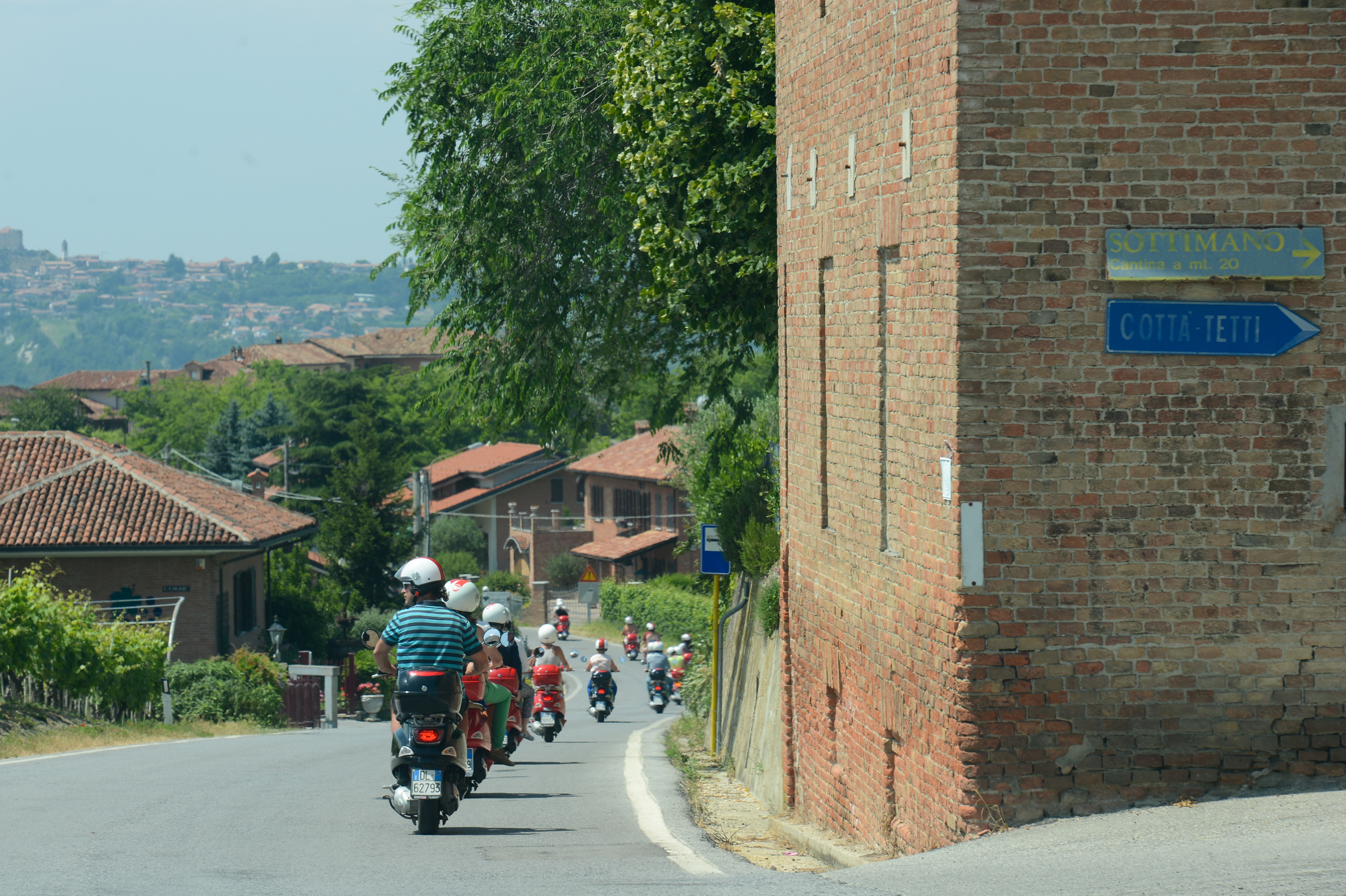 A group of people wearing helmets ride scooters down a winding road in a scenic countryside setting.