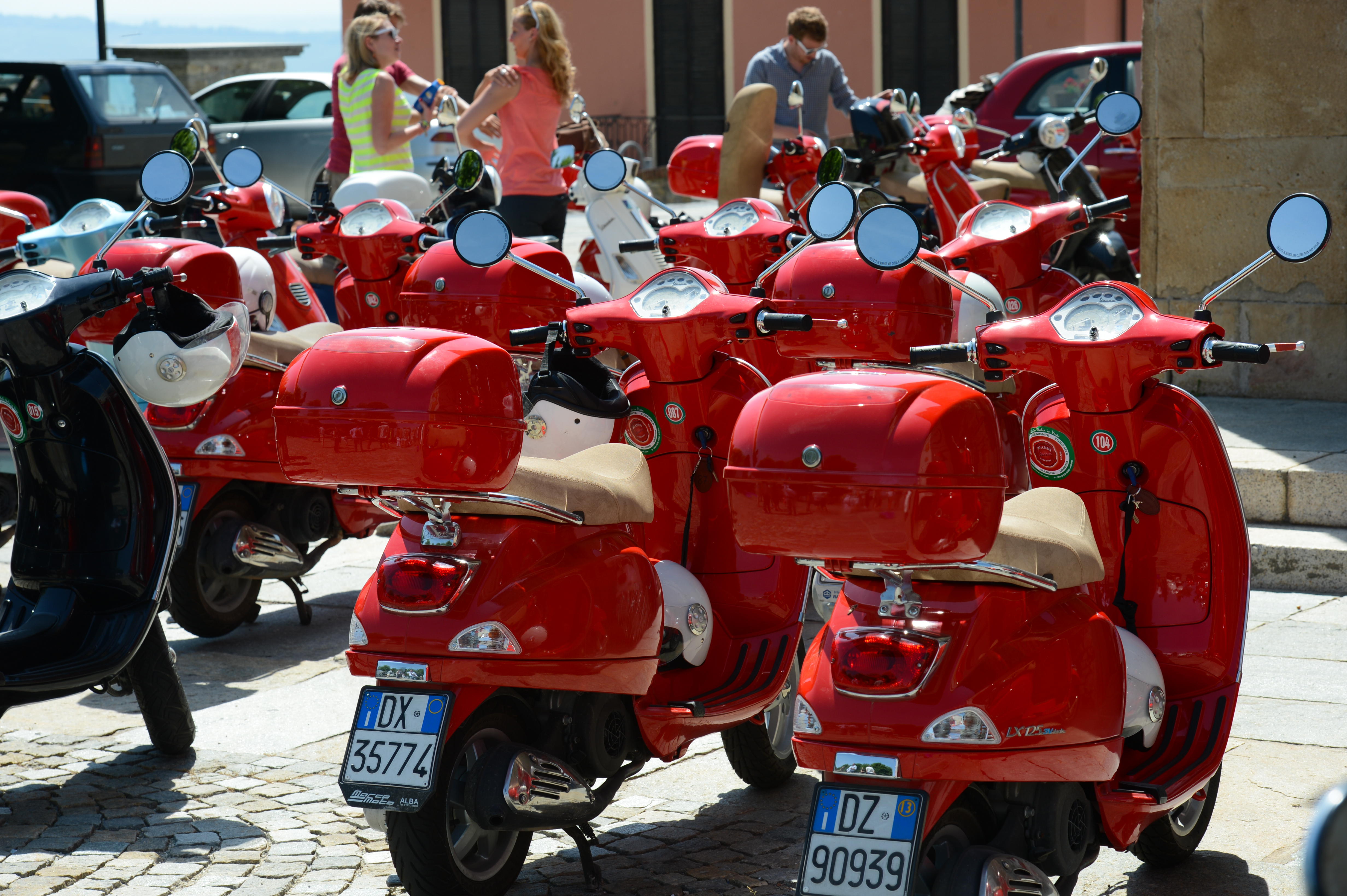 A row of red scooters parked closely together, with people standing nearby and one person adjusting a scooter.