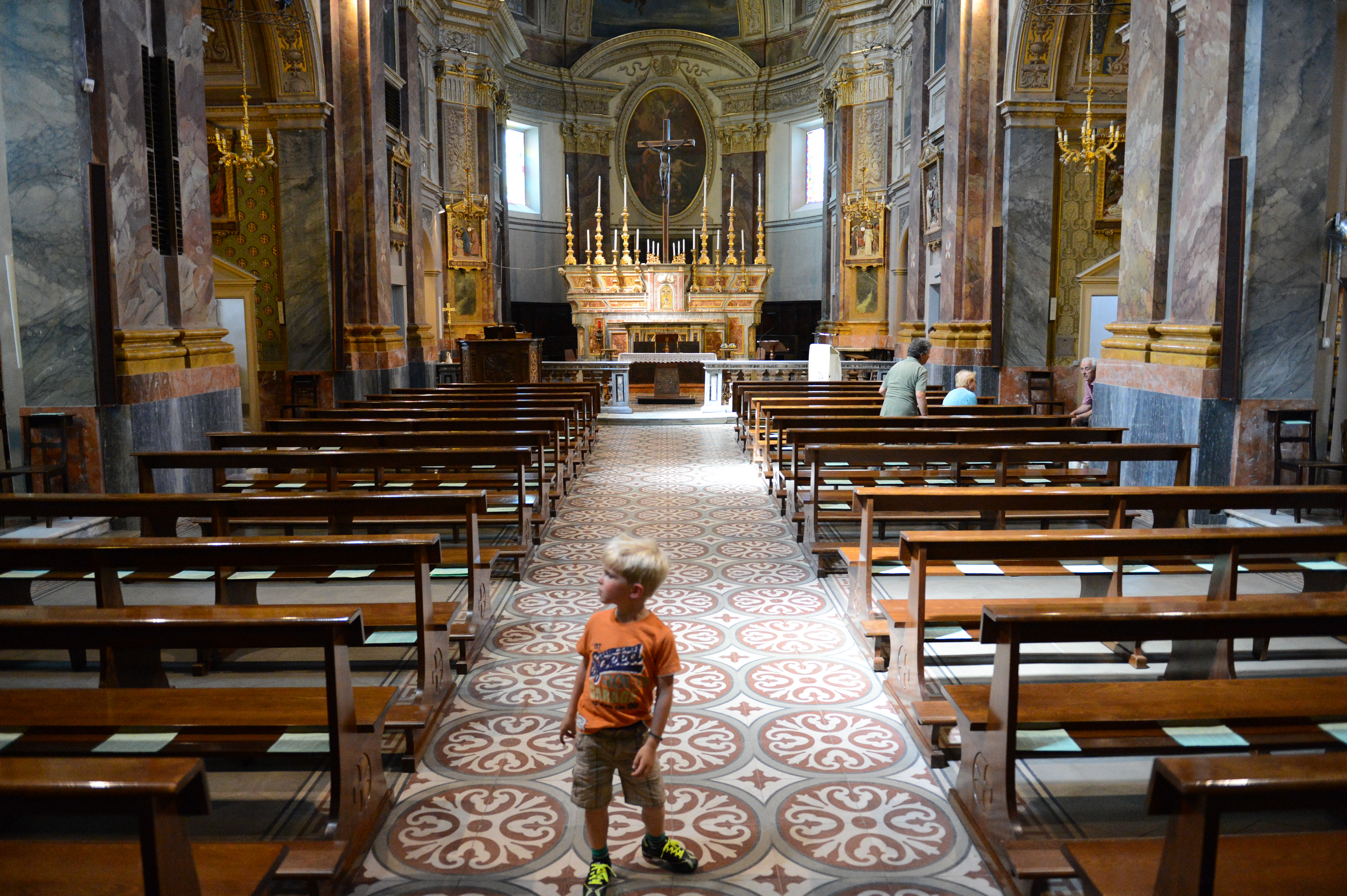 A young child walks down the aisle of a church, while a few people sit on the benches.