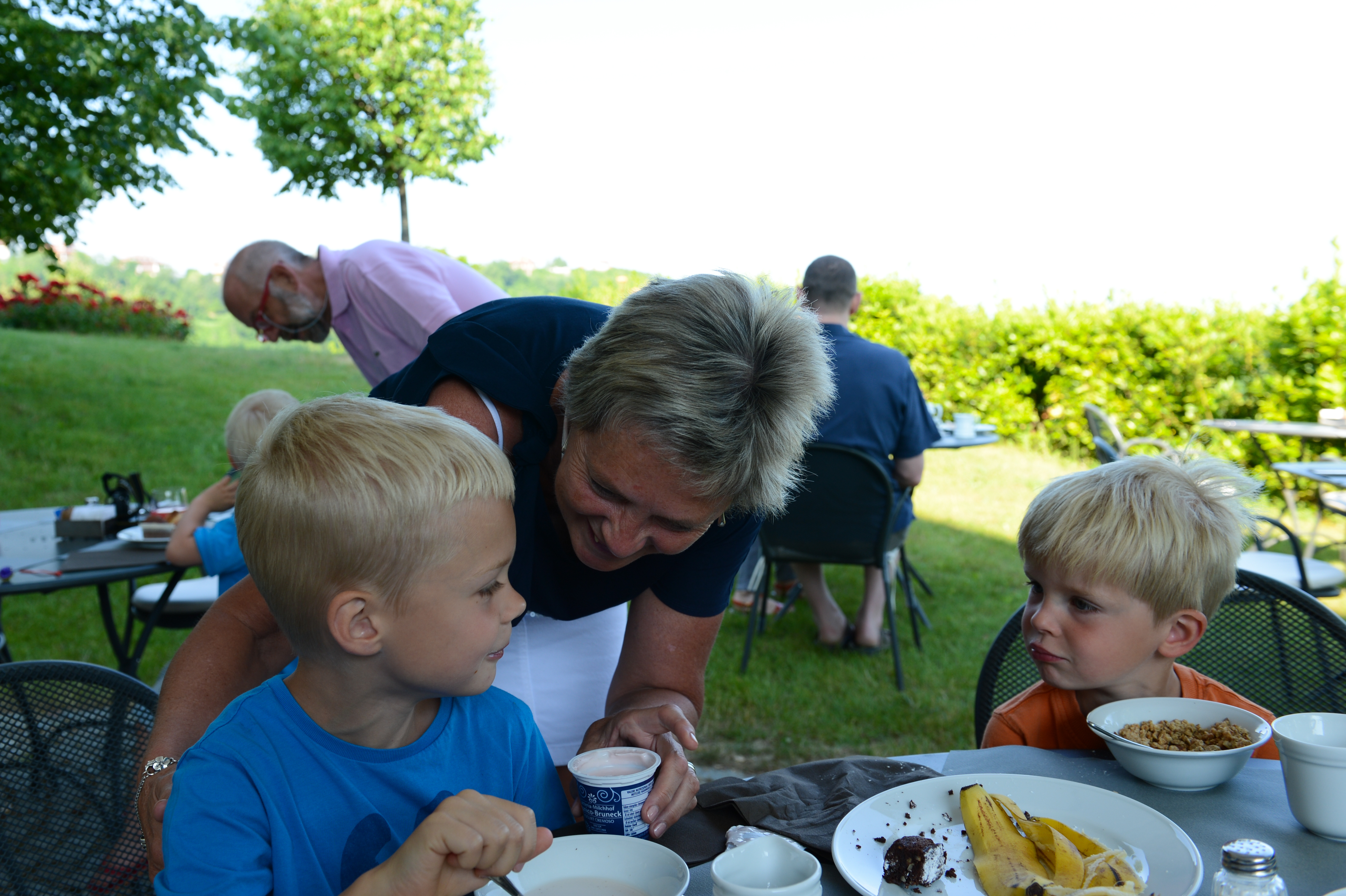 A woman leans in and smiles at a young boy while he eats at an outdoor gathering.
