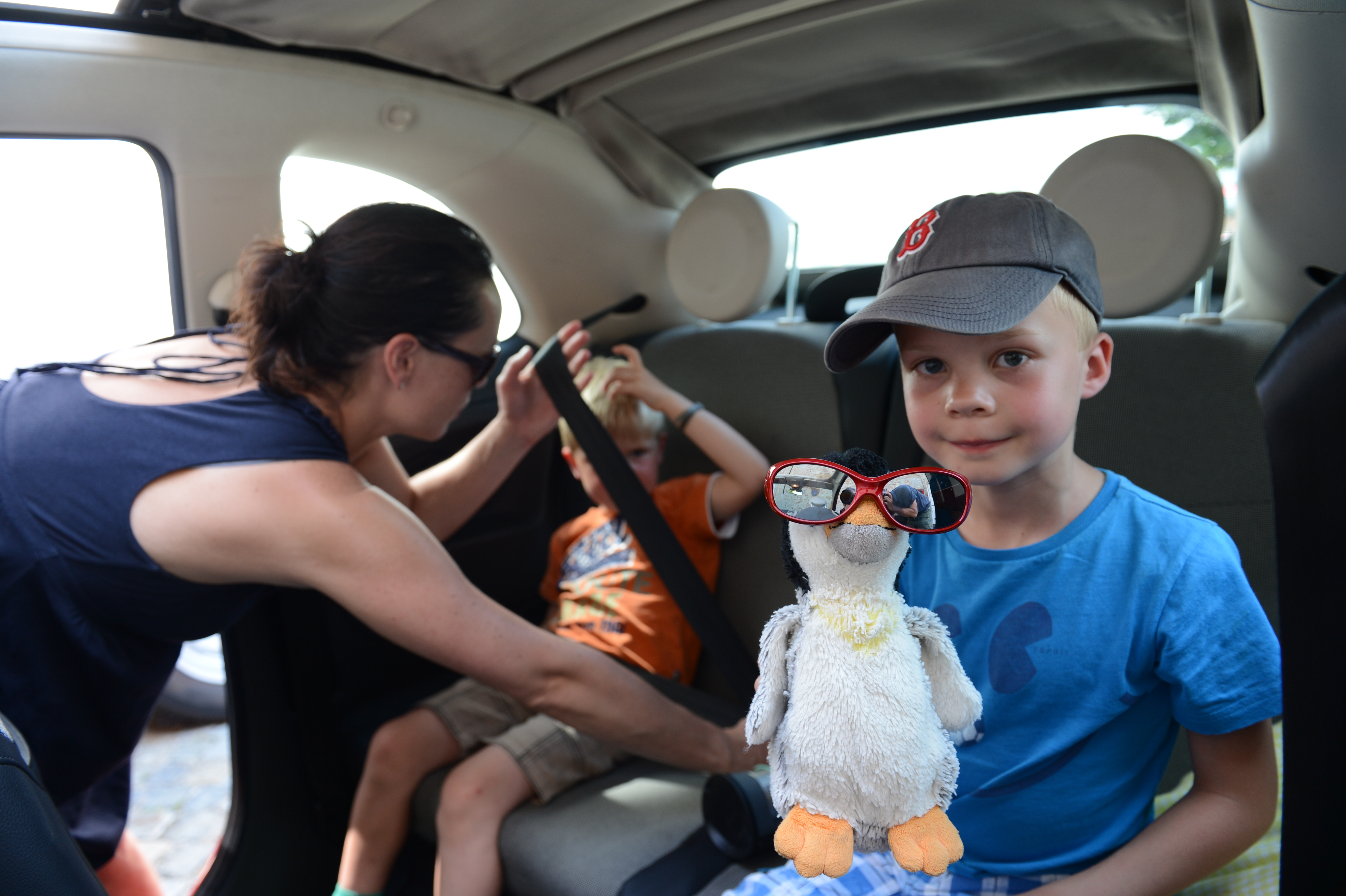 A woman helps a child buckle his seatbelt while another child holds a stuffed penguin wearing sunglasses.