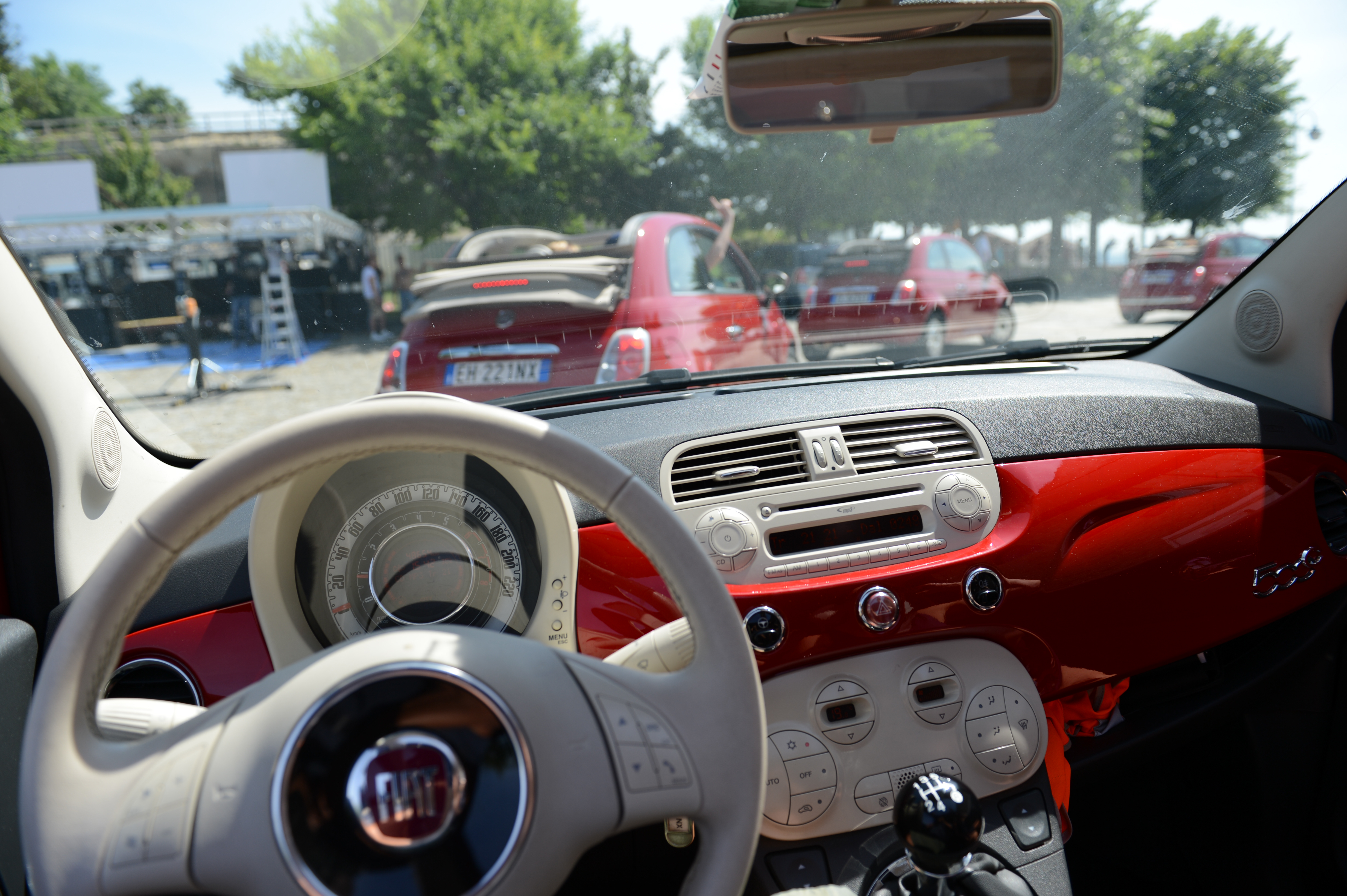 View from inside a red Fiat 500, looking out at similar cars parked ahead with people in the background.