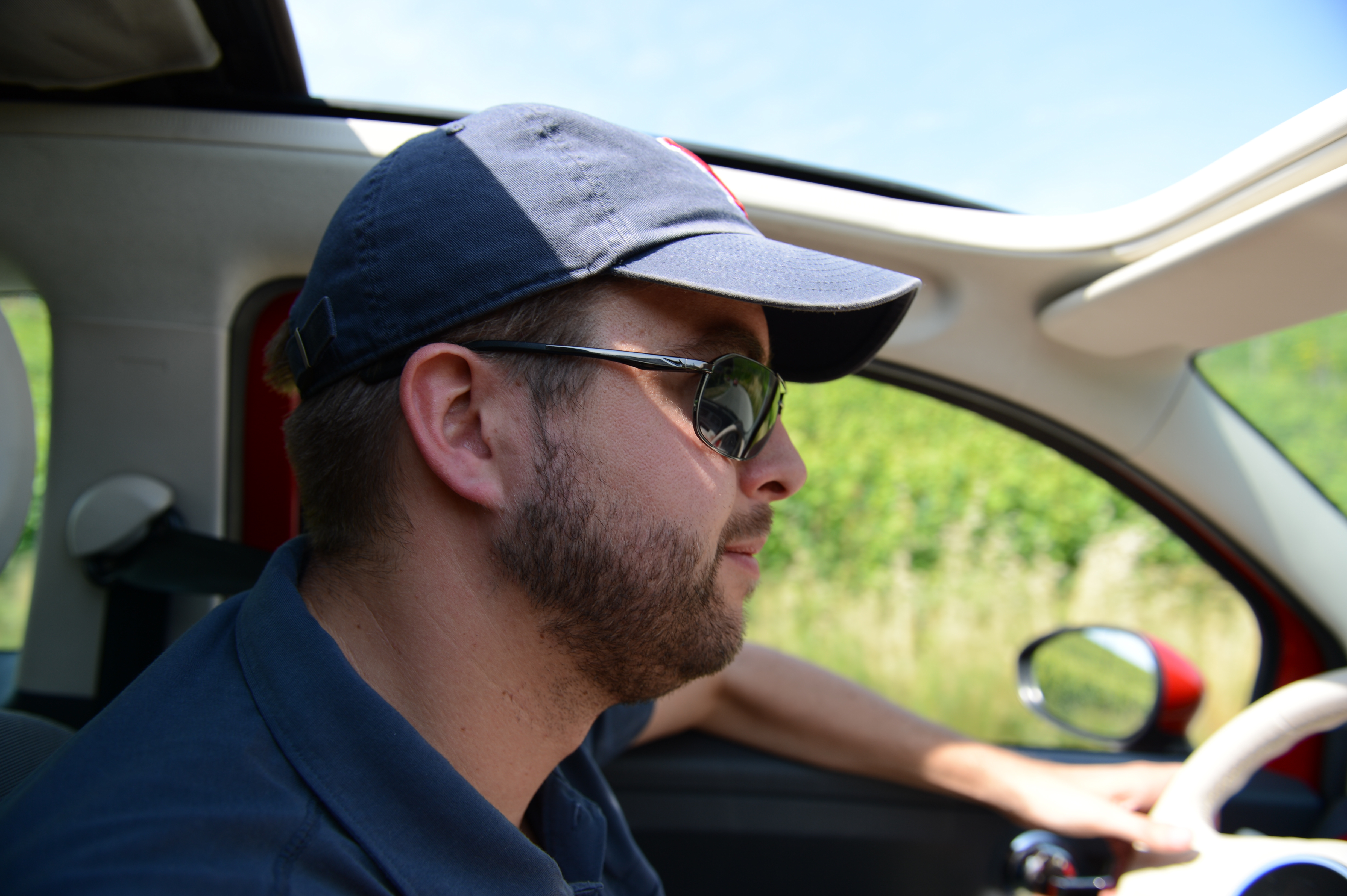 A man wearing sunglasses and a cap is driving a car with an open sunroof.