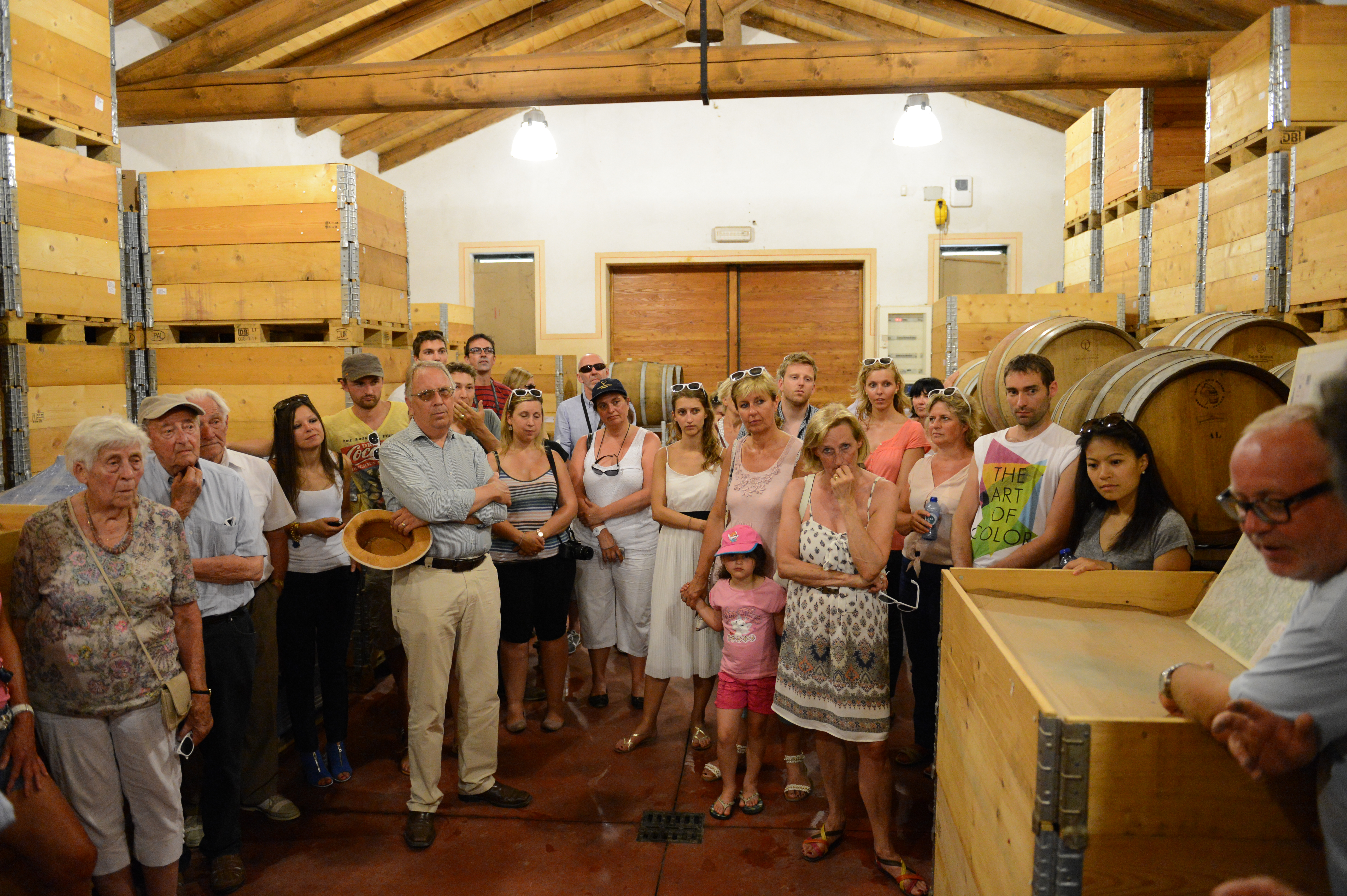 A group of wedding guests listens to a guide during a winery tour, standing among wooden barrels and crates.