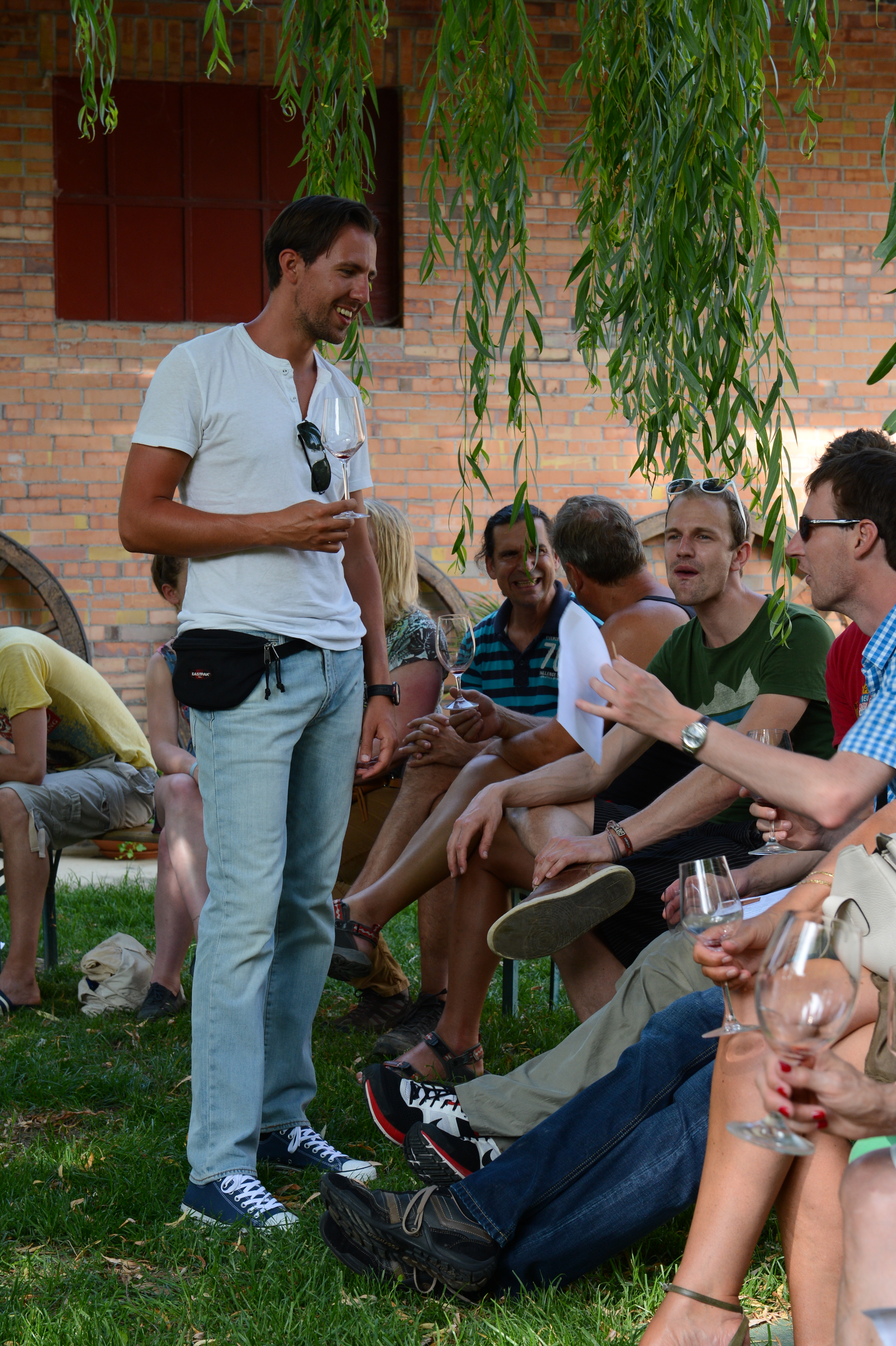 A man in a white shirt stands holding a wine glass, smiling and talking with seated guests at a wedding.