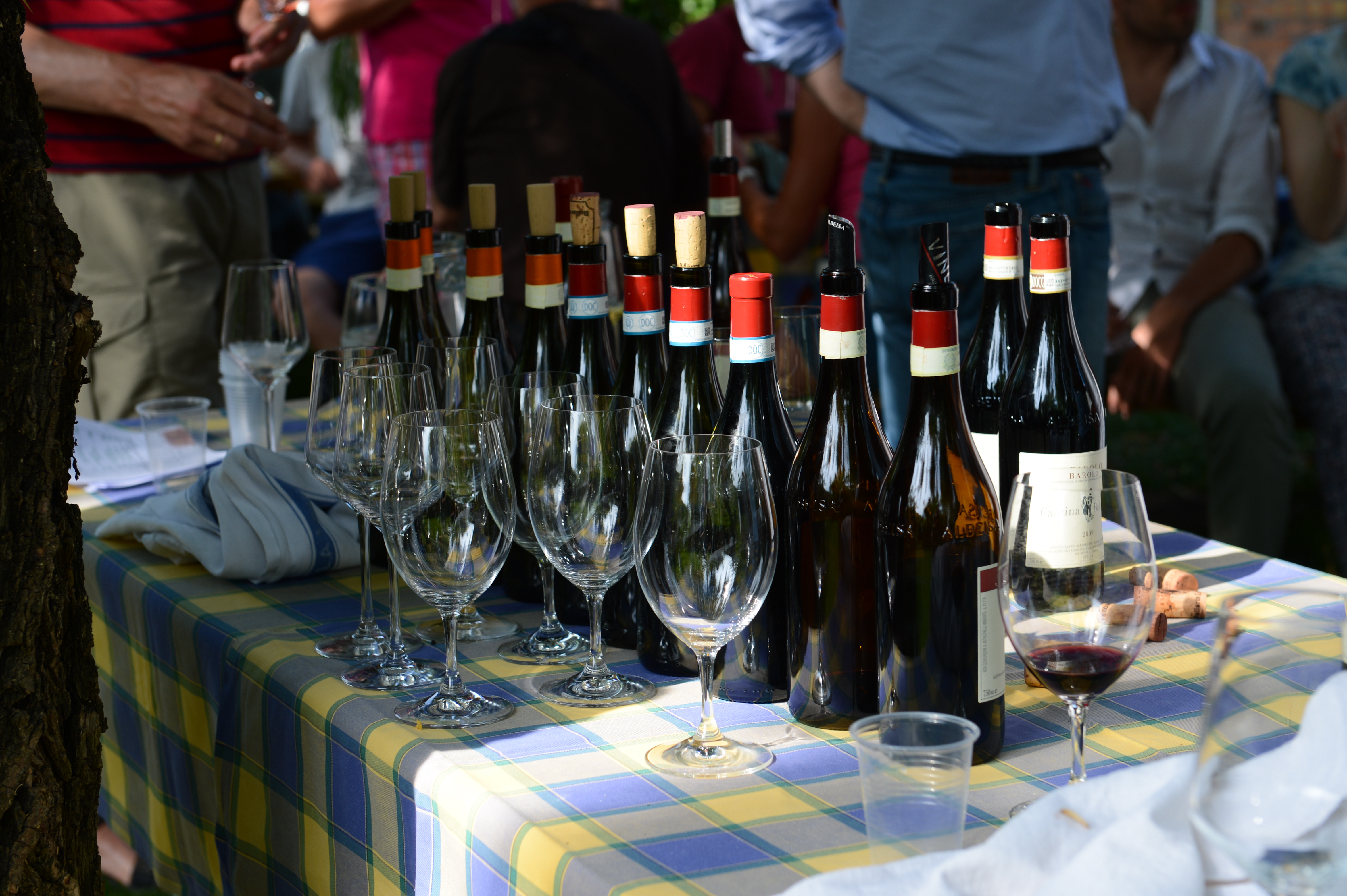 A table with wine bottles and empty glasses at an outdoor wedding reception, with guests standing in the background.