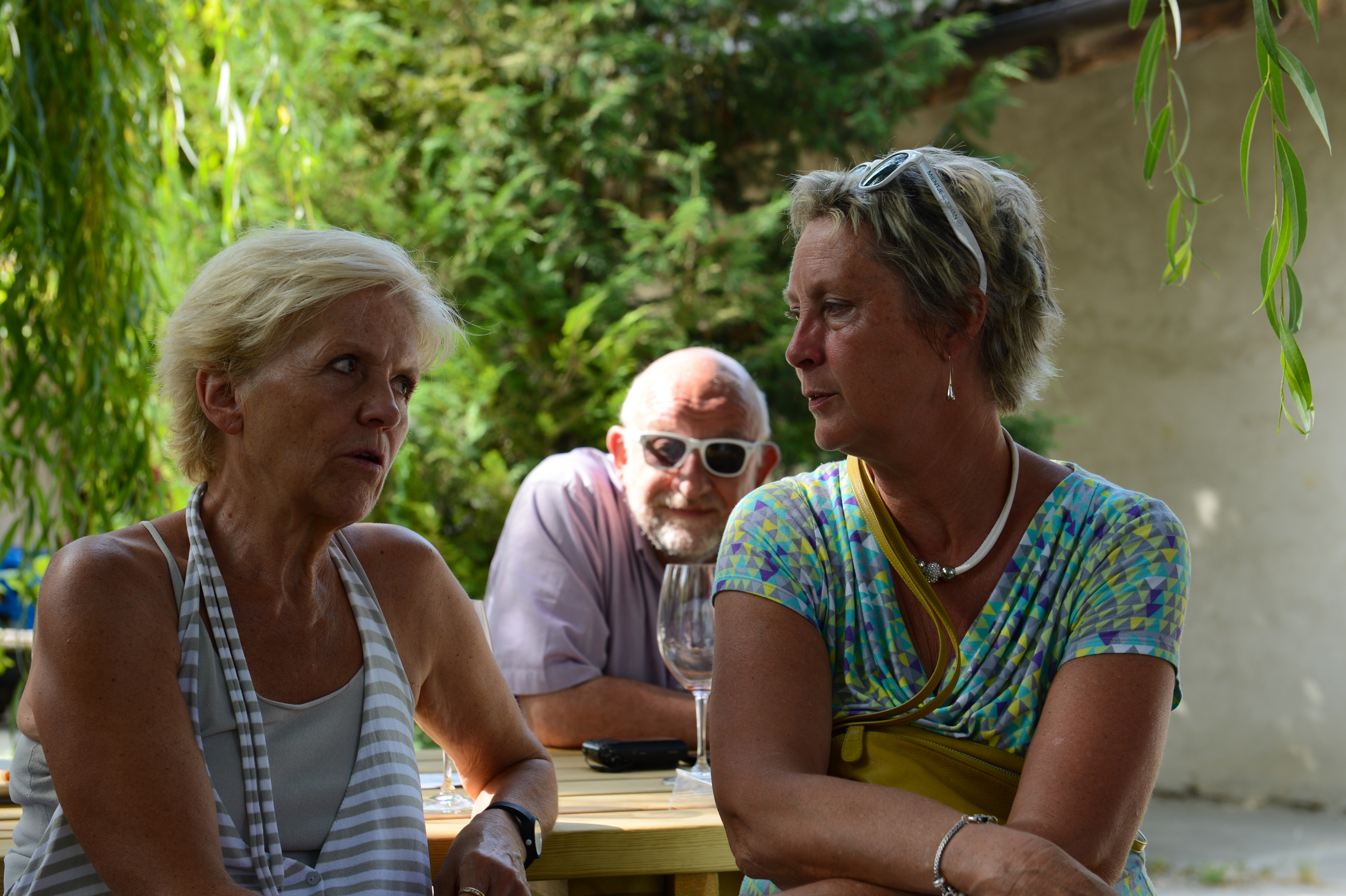 Two women sit at a table engaged in conversation, while a man in sunglasses watches from behind with a drink.