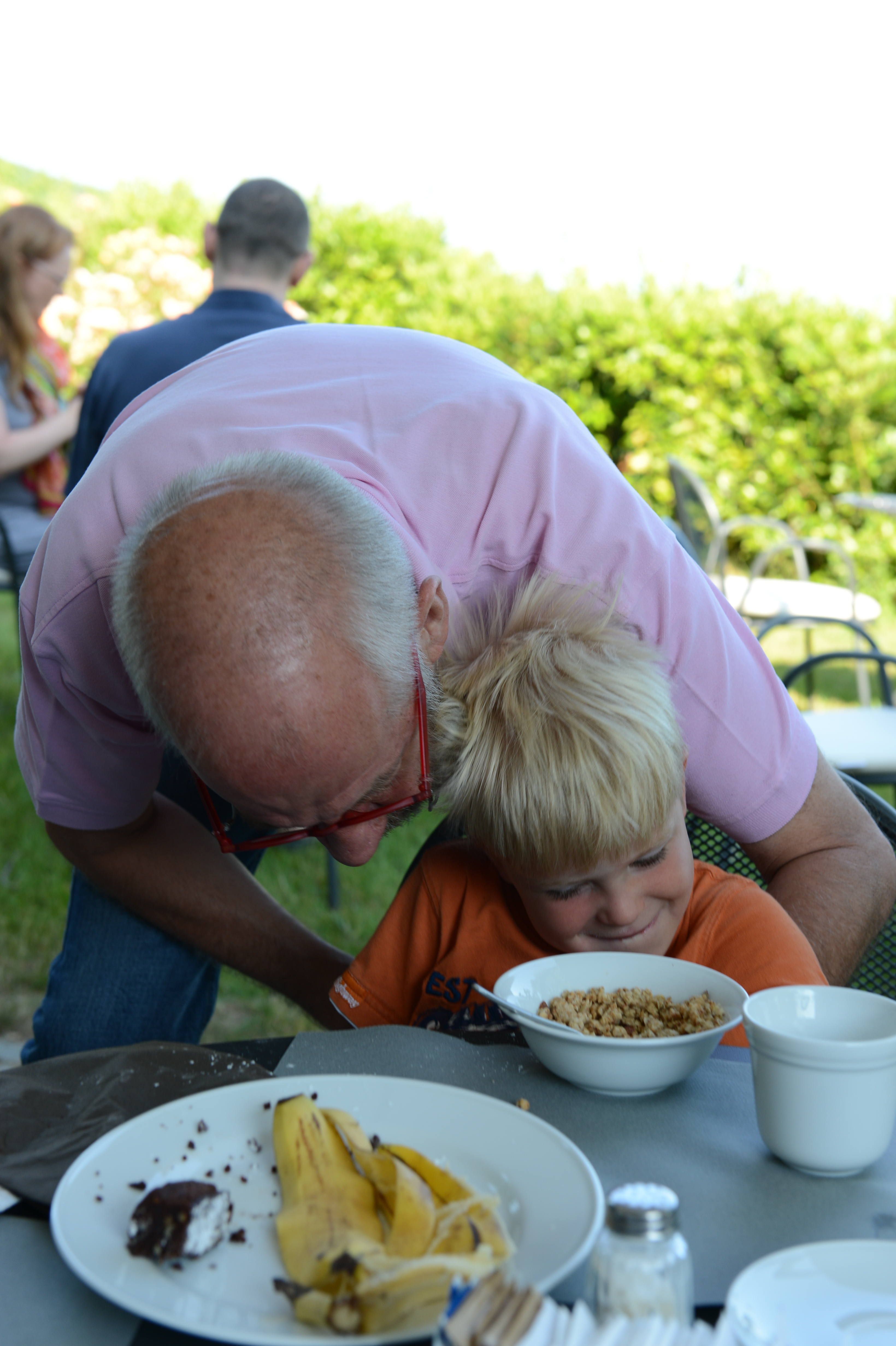 An older man in a pink shirt leans down playfully toward a smiling young boy at a table with food.