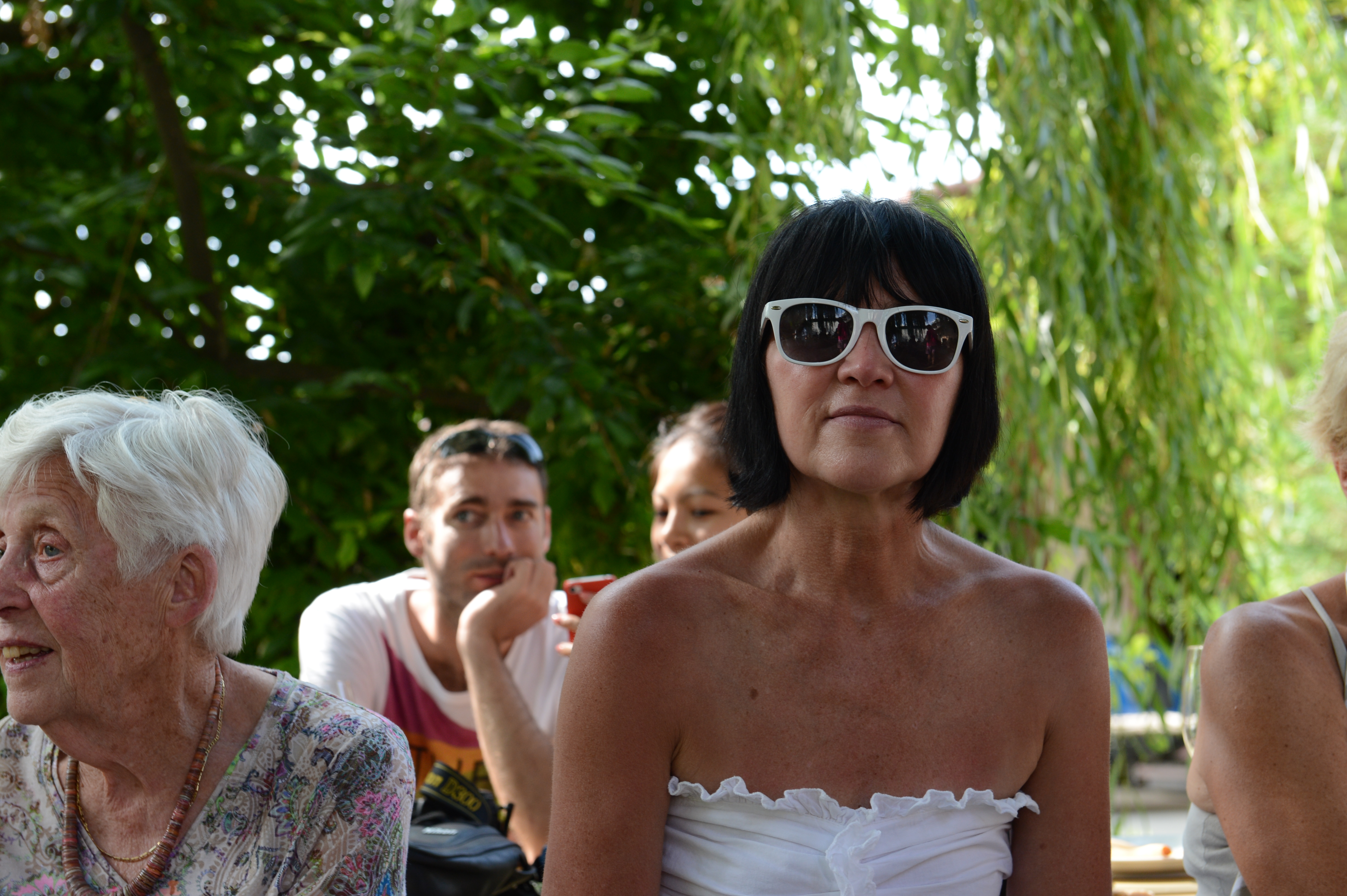 A woman in white sunglasses sits at an outdoor wedding gathering, with other guests in the background.