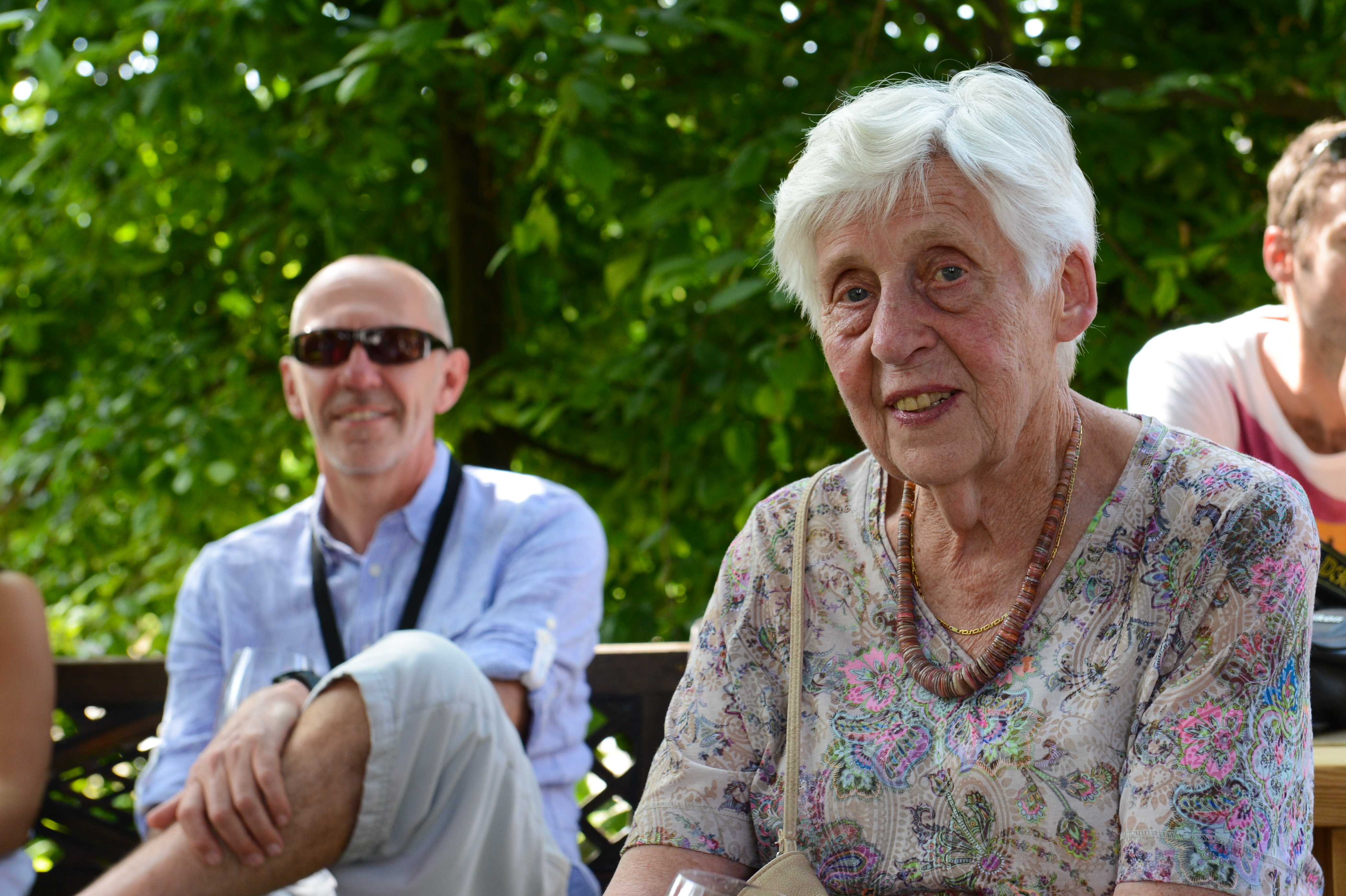 An elderly woman smiles at the camera while seated outdoors, with a man in sunglasses sitting behind her.