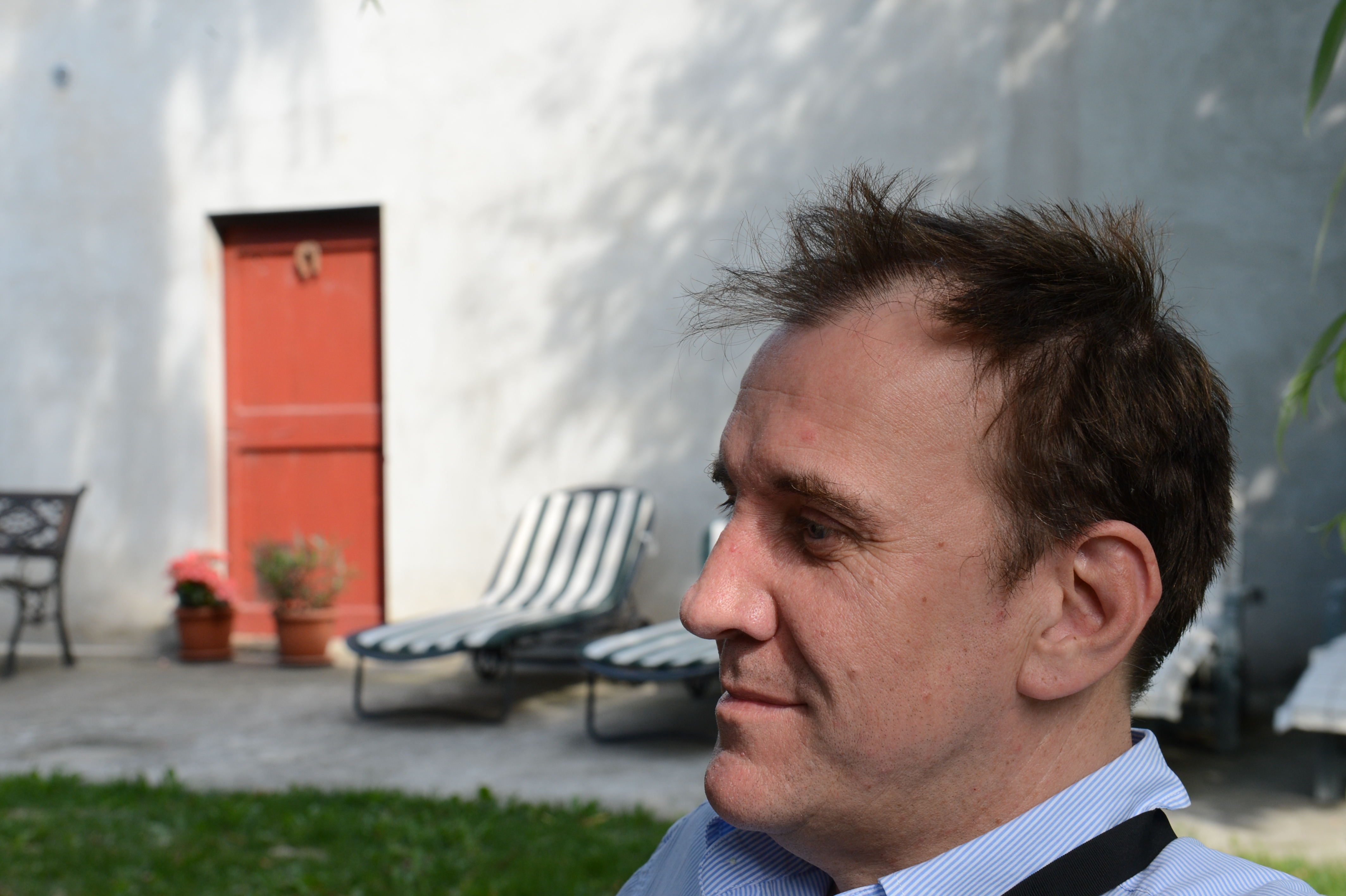 A man in a blue shirt sits outdoors, looking to the side with a relaxed expression.