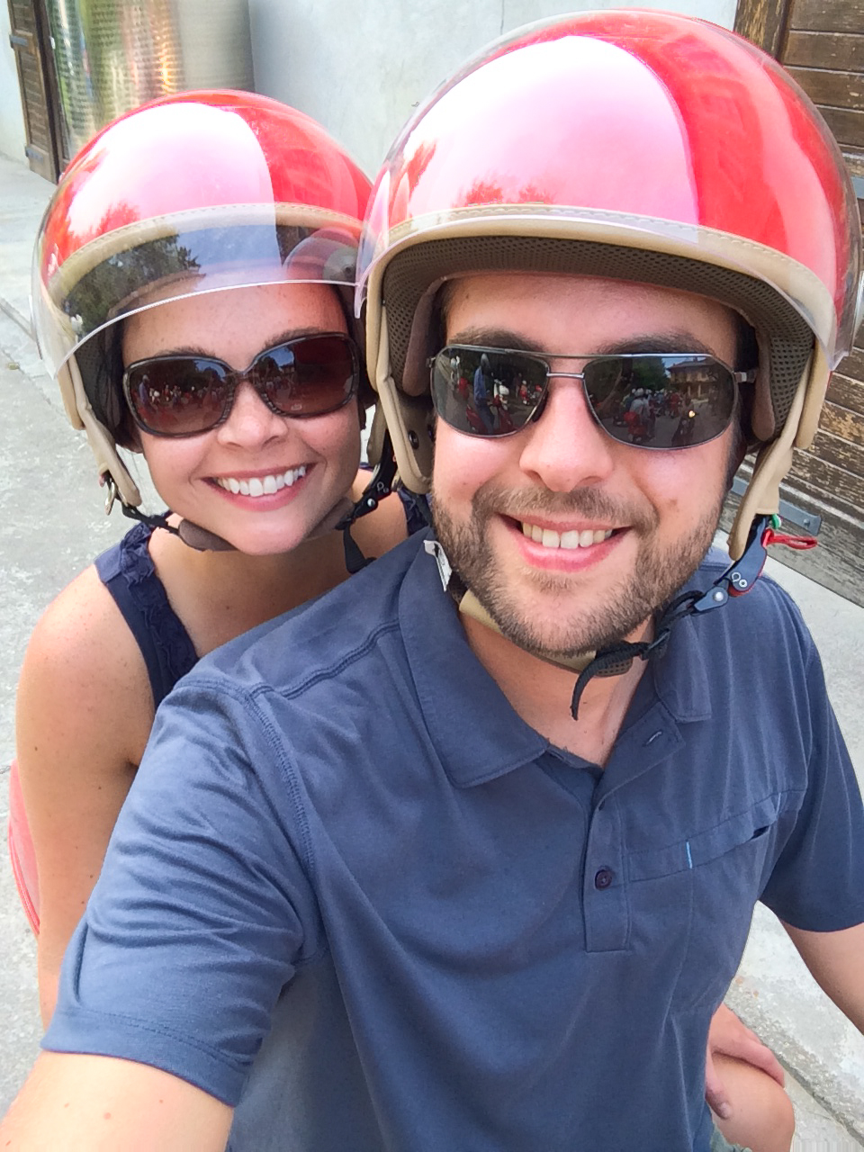 A smiling couple wearing helmets and sunglasses takes a selfie while sitting on a motorbike.