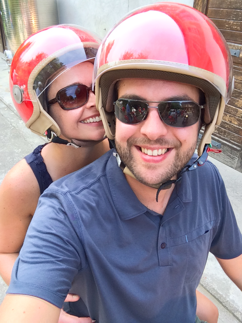 A smiling couple wearing red helmets and sunglasses pose for a selfie while sitting on a scooter.