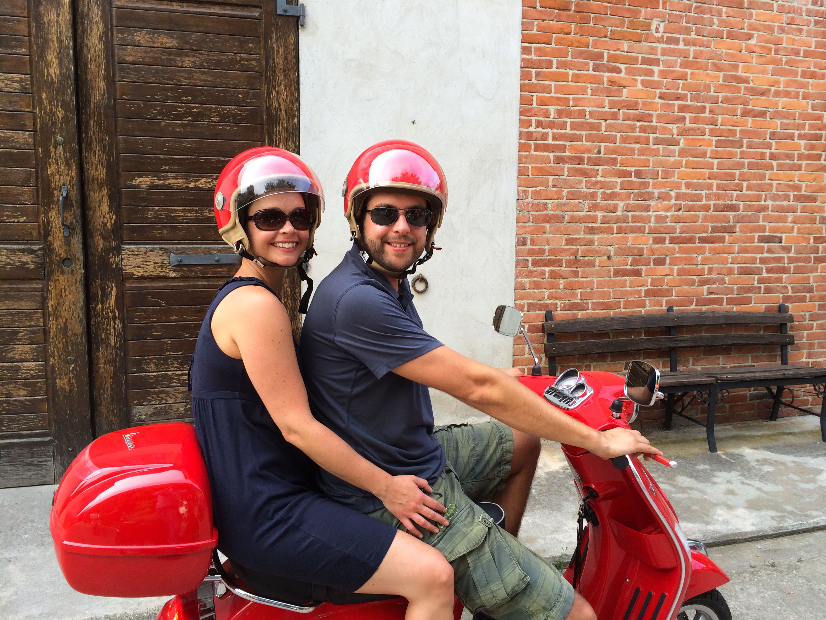 A smiling couple wearing helmets sits on a red scooter, with the man holding the handlebars.