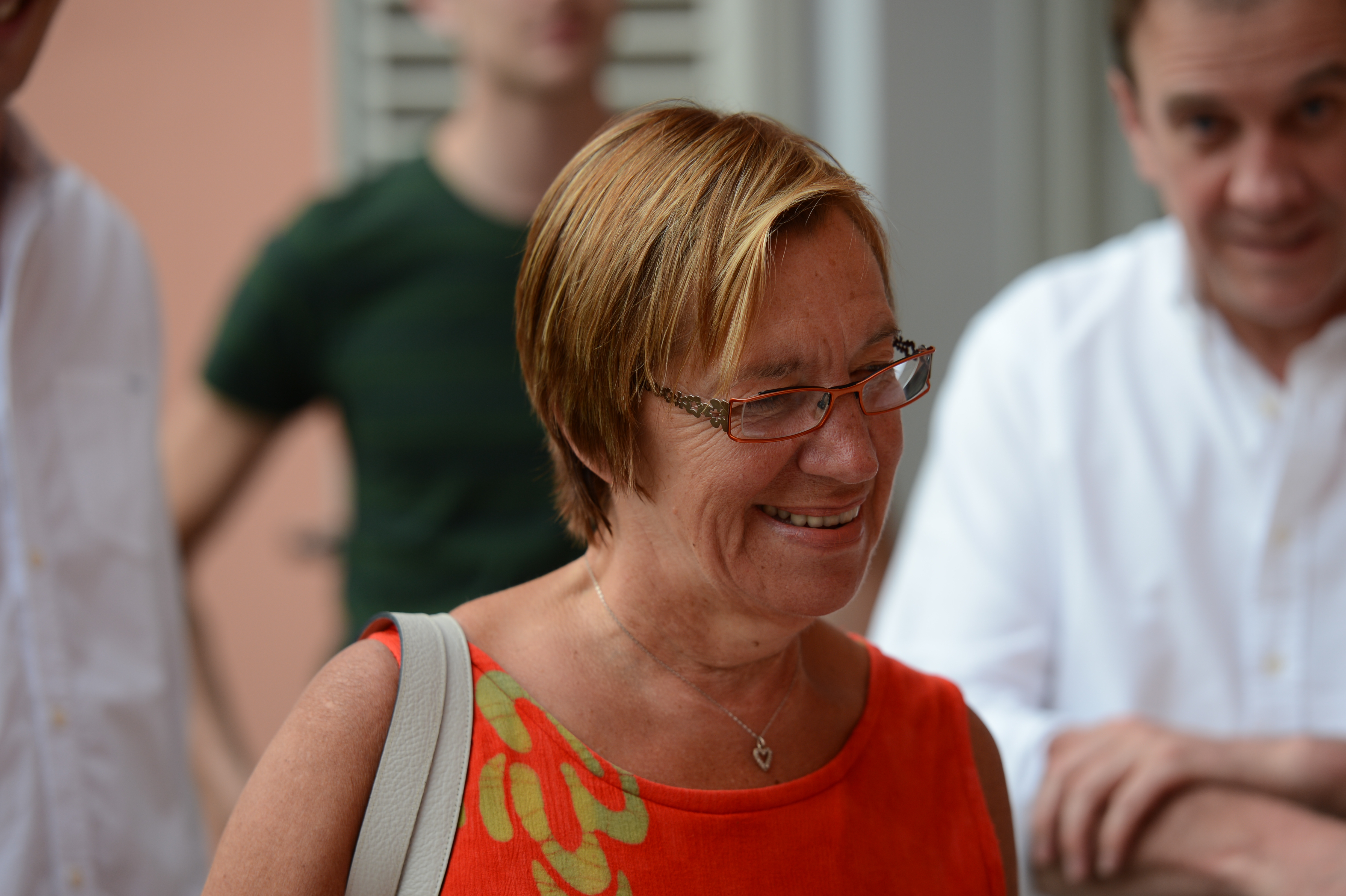 A woman in glasses and an orange dress smiles while talking at a wedding gathering.