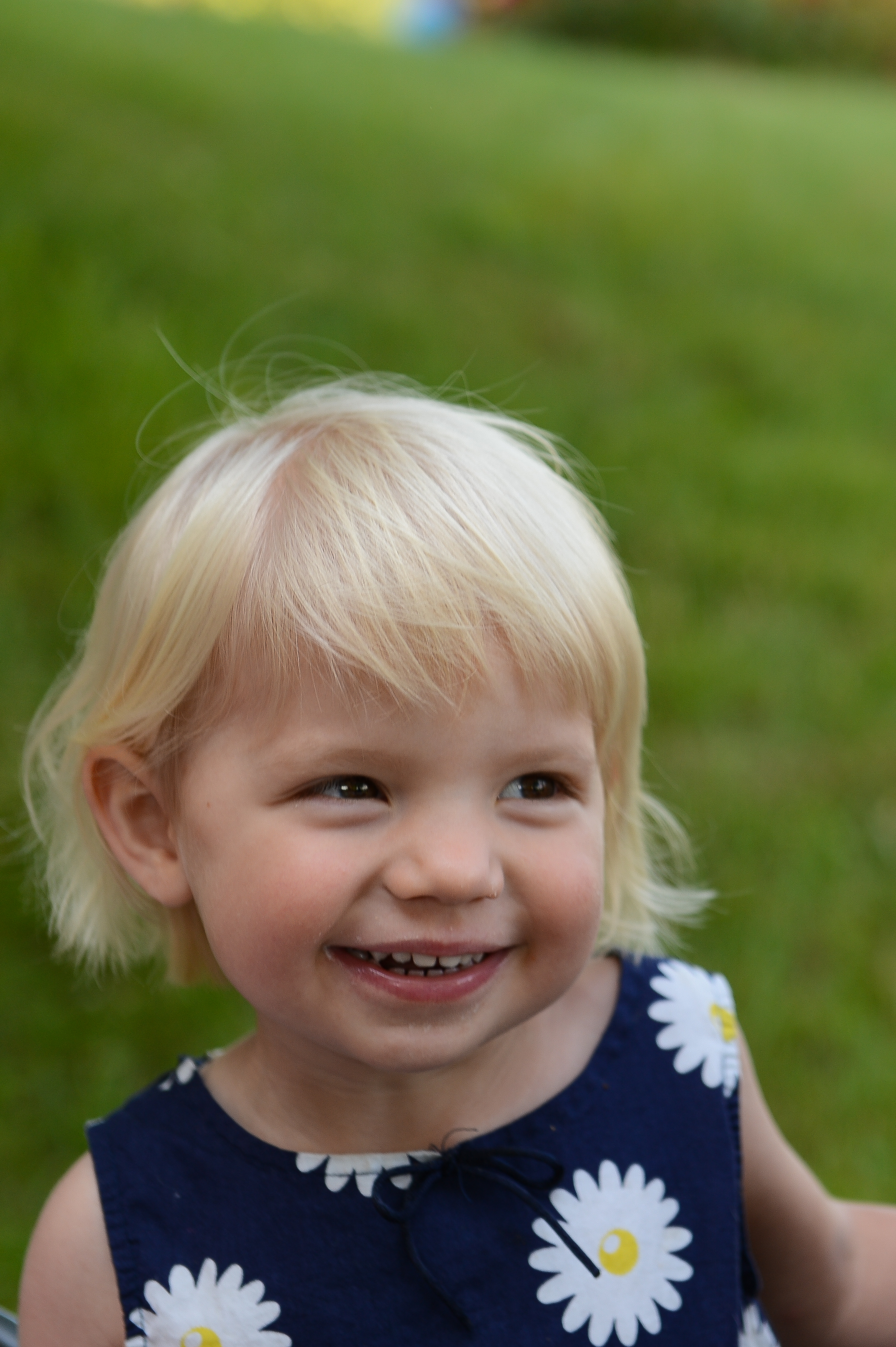 A smiling young child in a blue dress with white flowers, looking slightly to the side at an outdoor wedding.