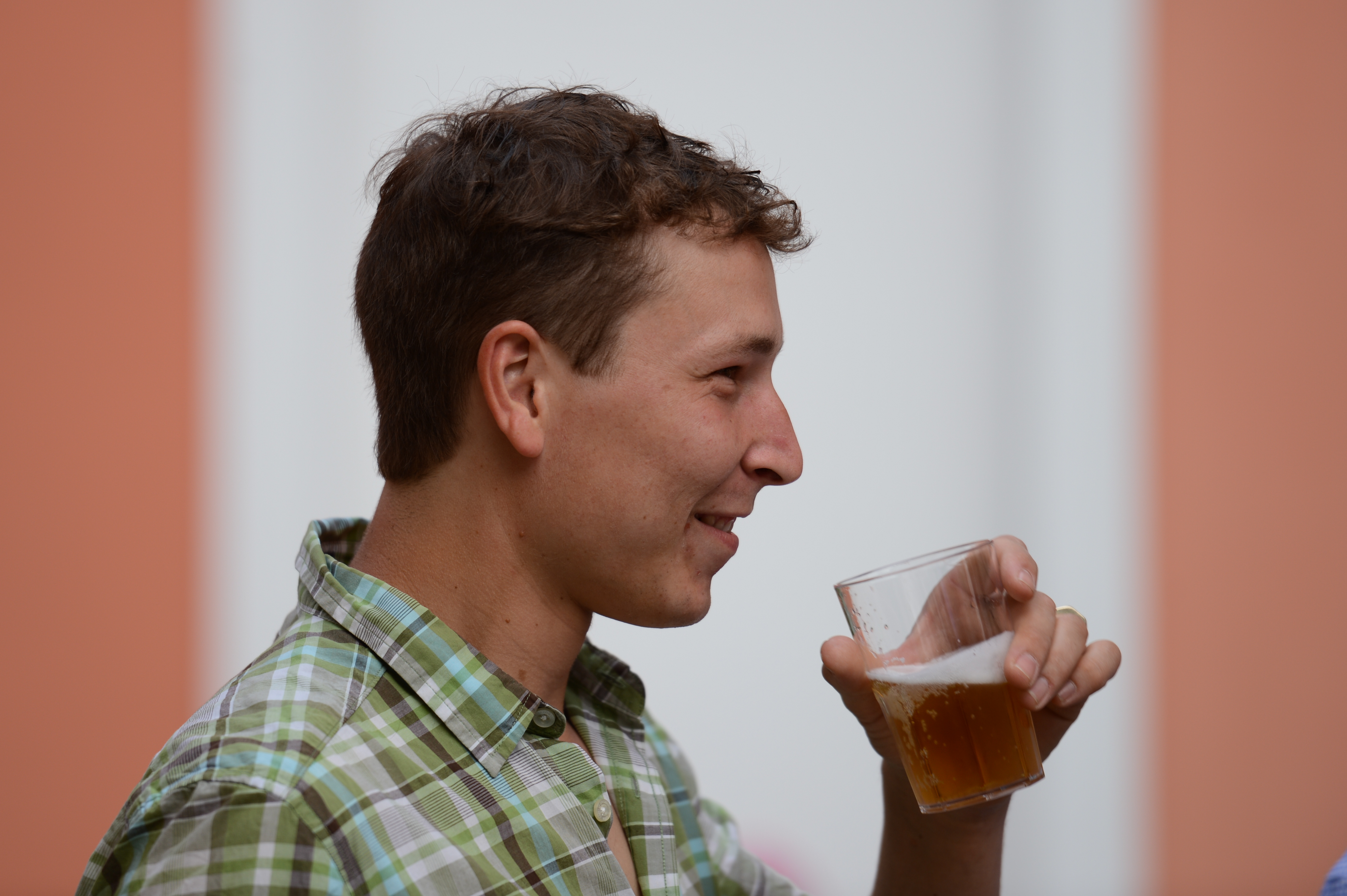 A man in a plaid shirt holds a glass of beer and smiles while looking to the side.