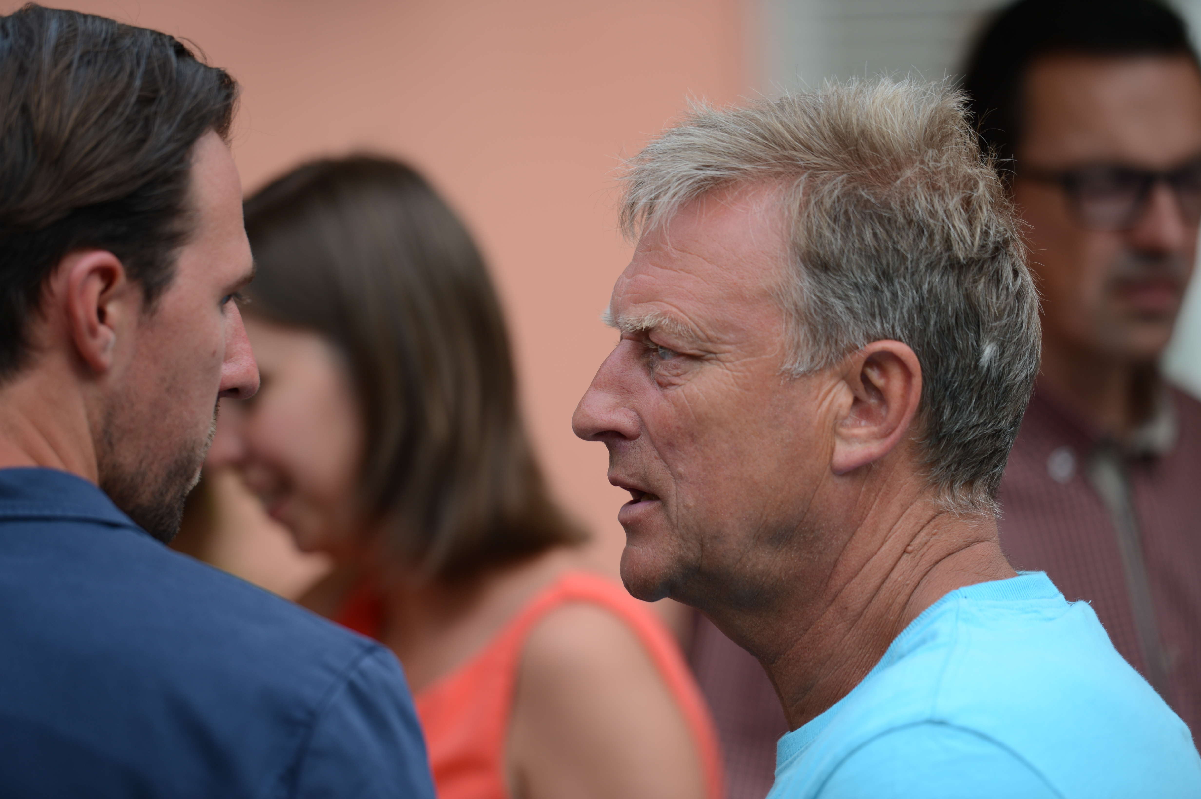 Two men engaged in conversation at a wedding, with other guests visible in the background.