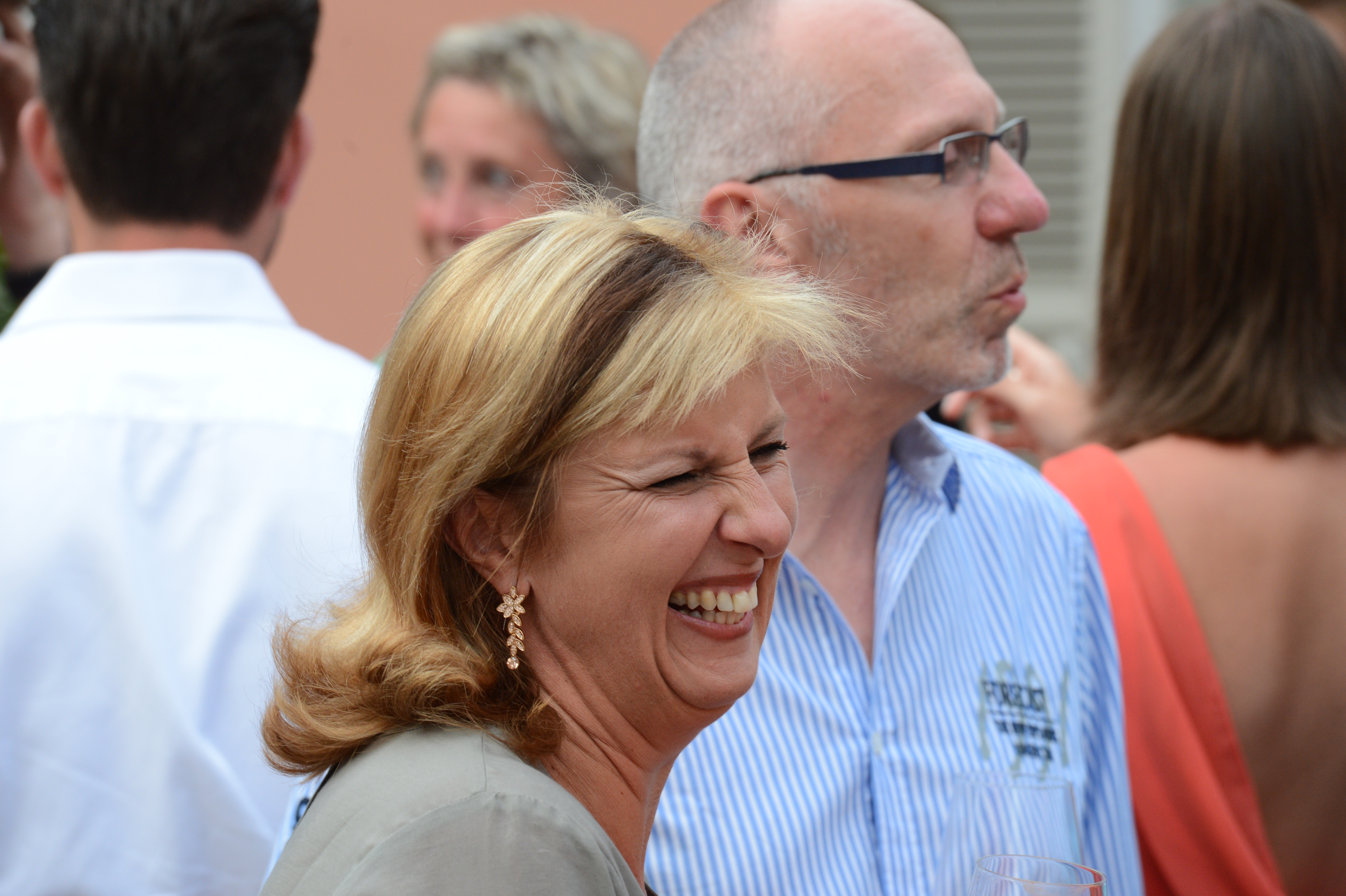 A woman with blonde hair laughs joyfully at a wedding reception, surrounded by other guests in conversation.