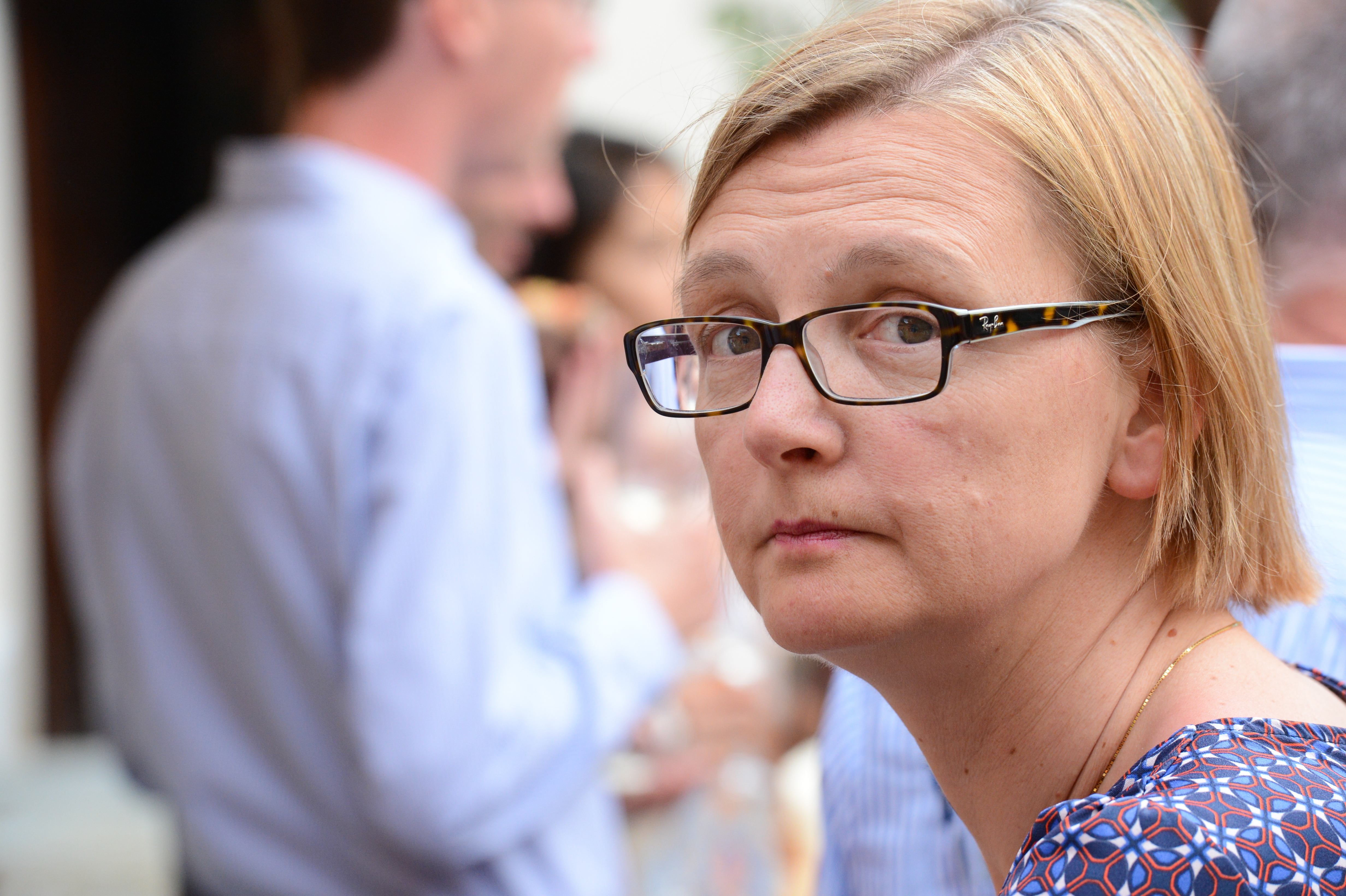 A woman with glasses looks toward the camera at a wedding reception, with guests socializing in the background.