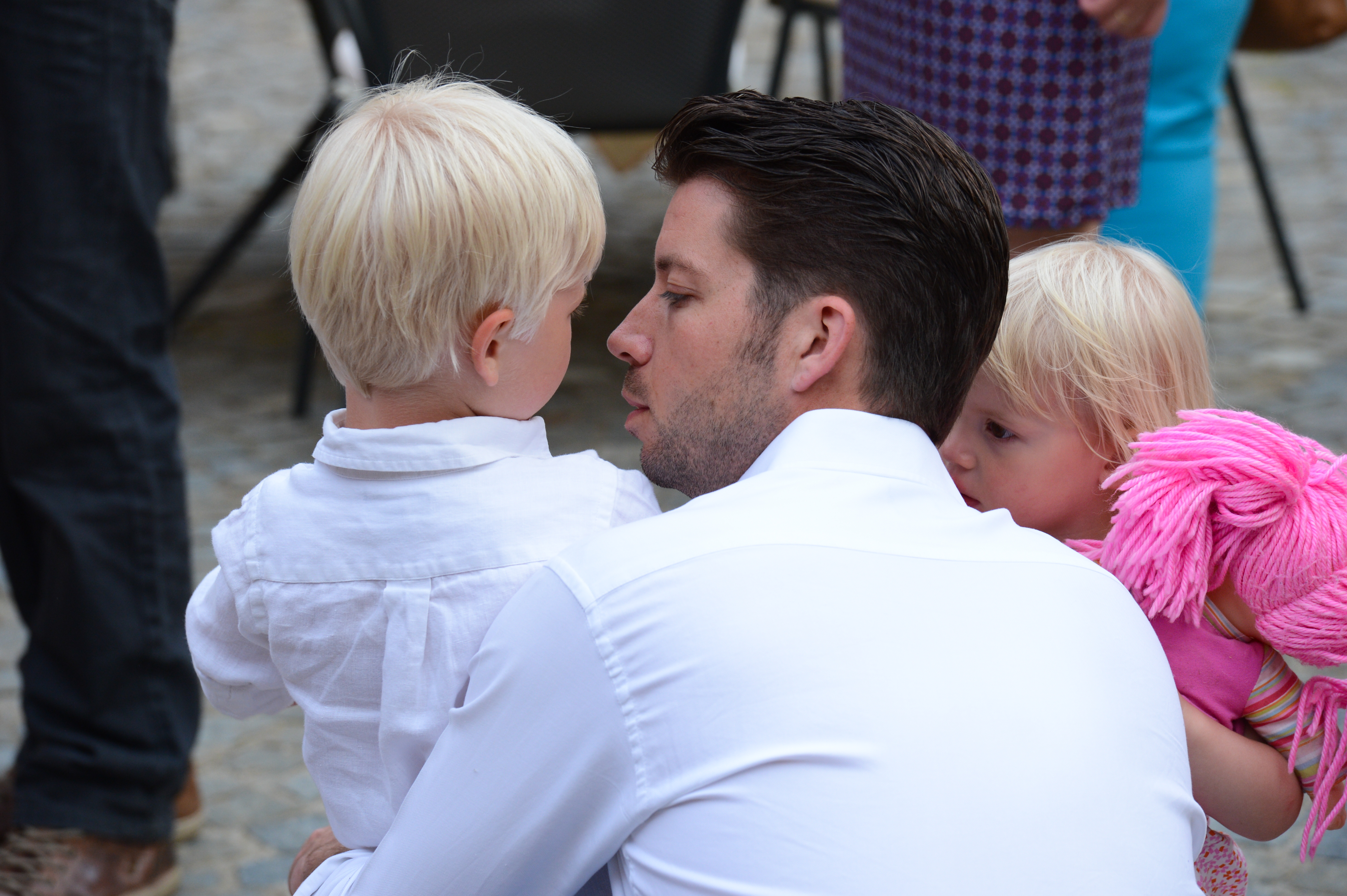 A man in a white shirt crouches, holding a young boy while a little girl clings to him with a doll.