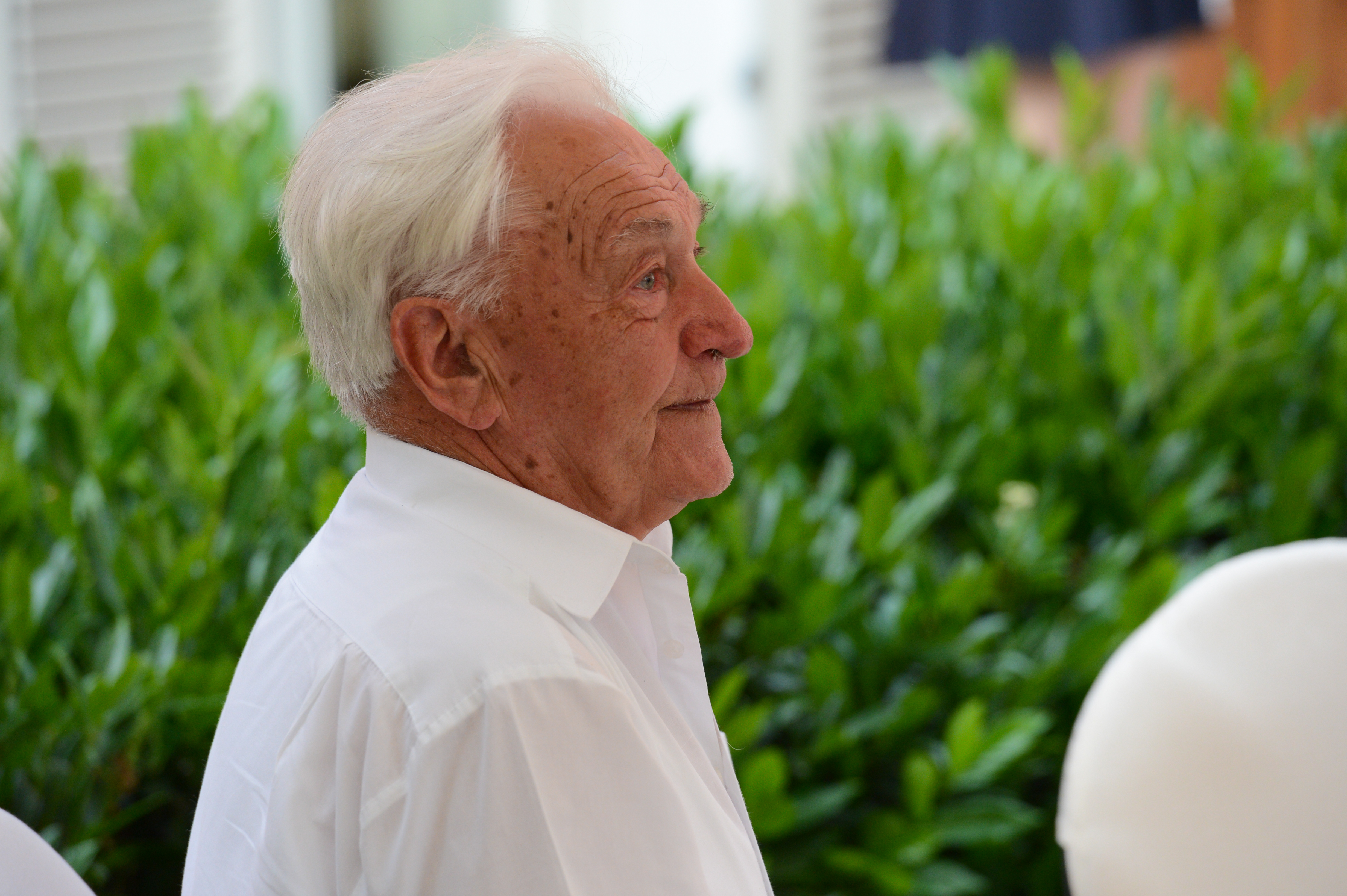 An elderly man in a white shirt sits outdoors, attentively watching something at a wedding.