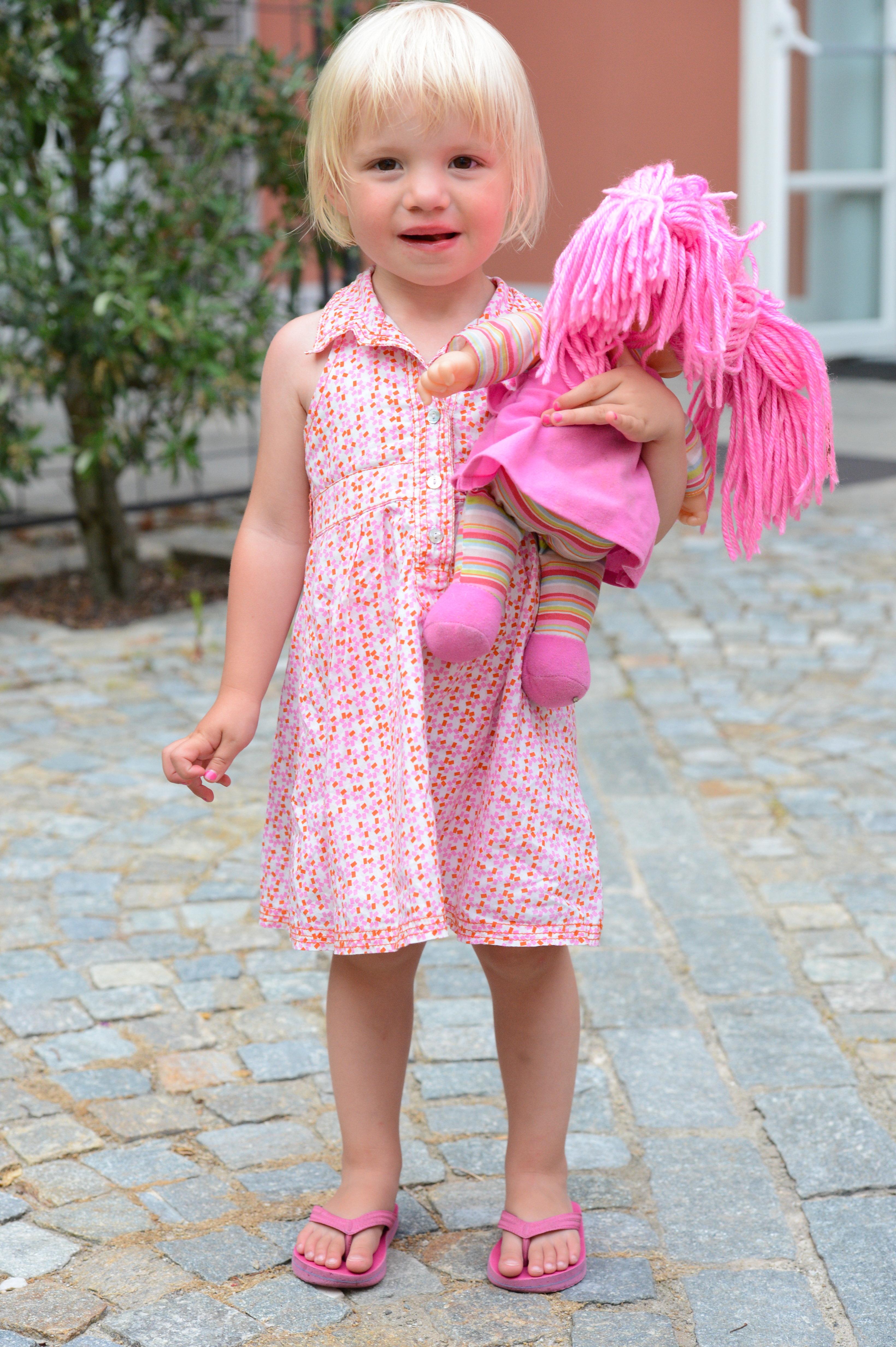 A young child in a pink dress holds a doll with pink yarn hair, standing on a cobblestone path.