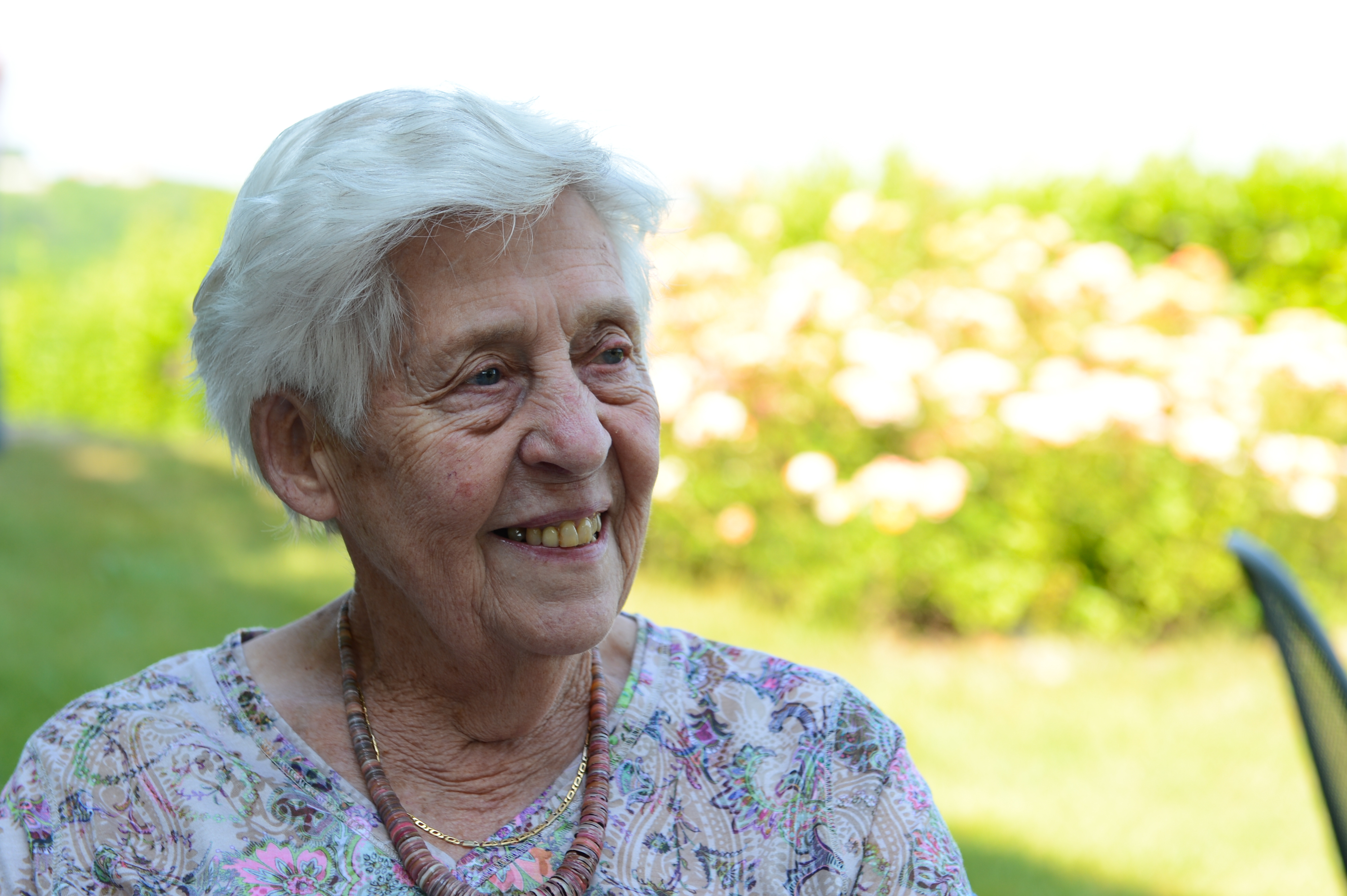 An elderly woman with short white hair smiles while sitting outdoors at a wedding.