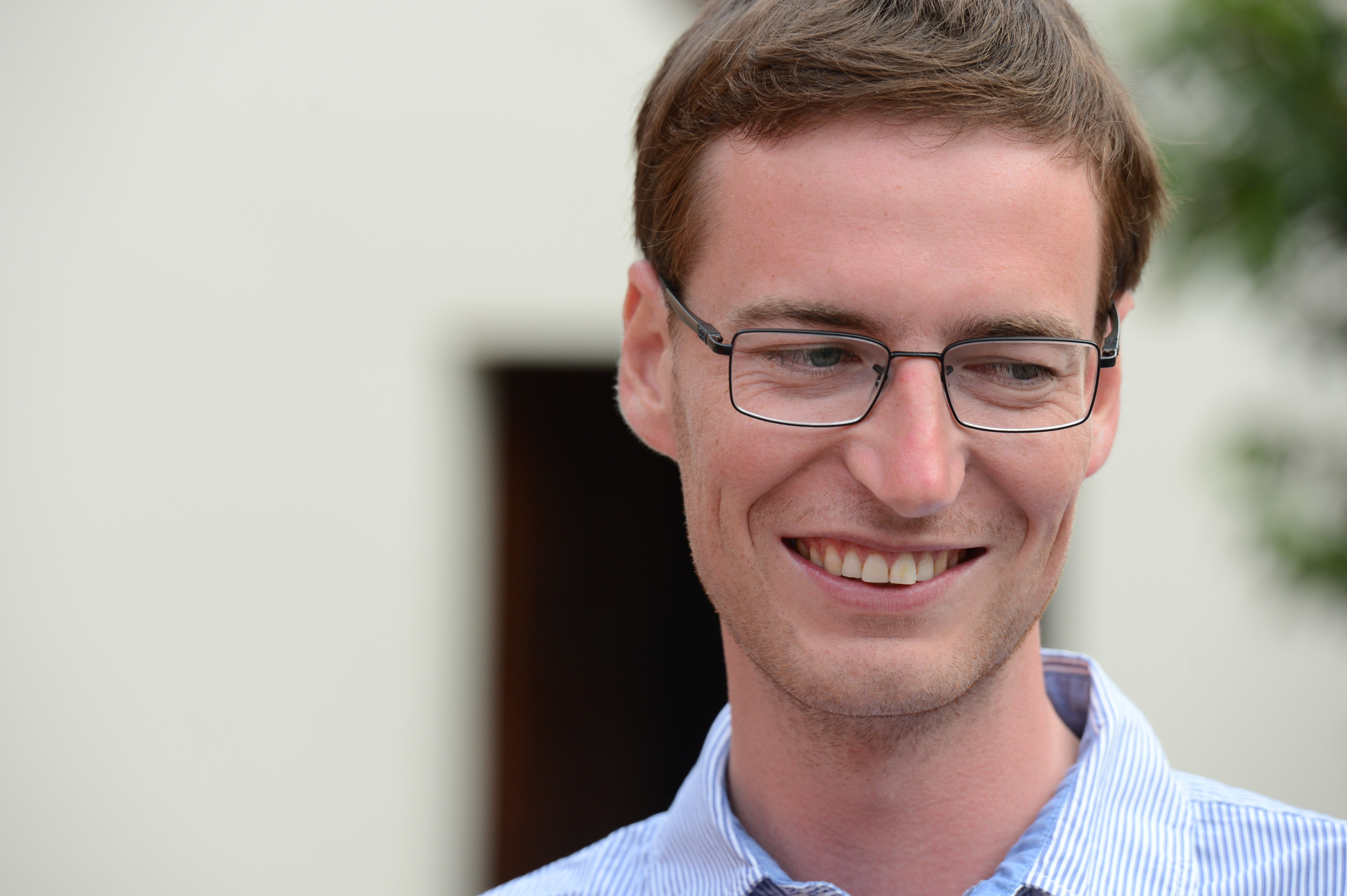 A man wearing glasses and a striped shirt smiles while looking to the side at a wedding.