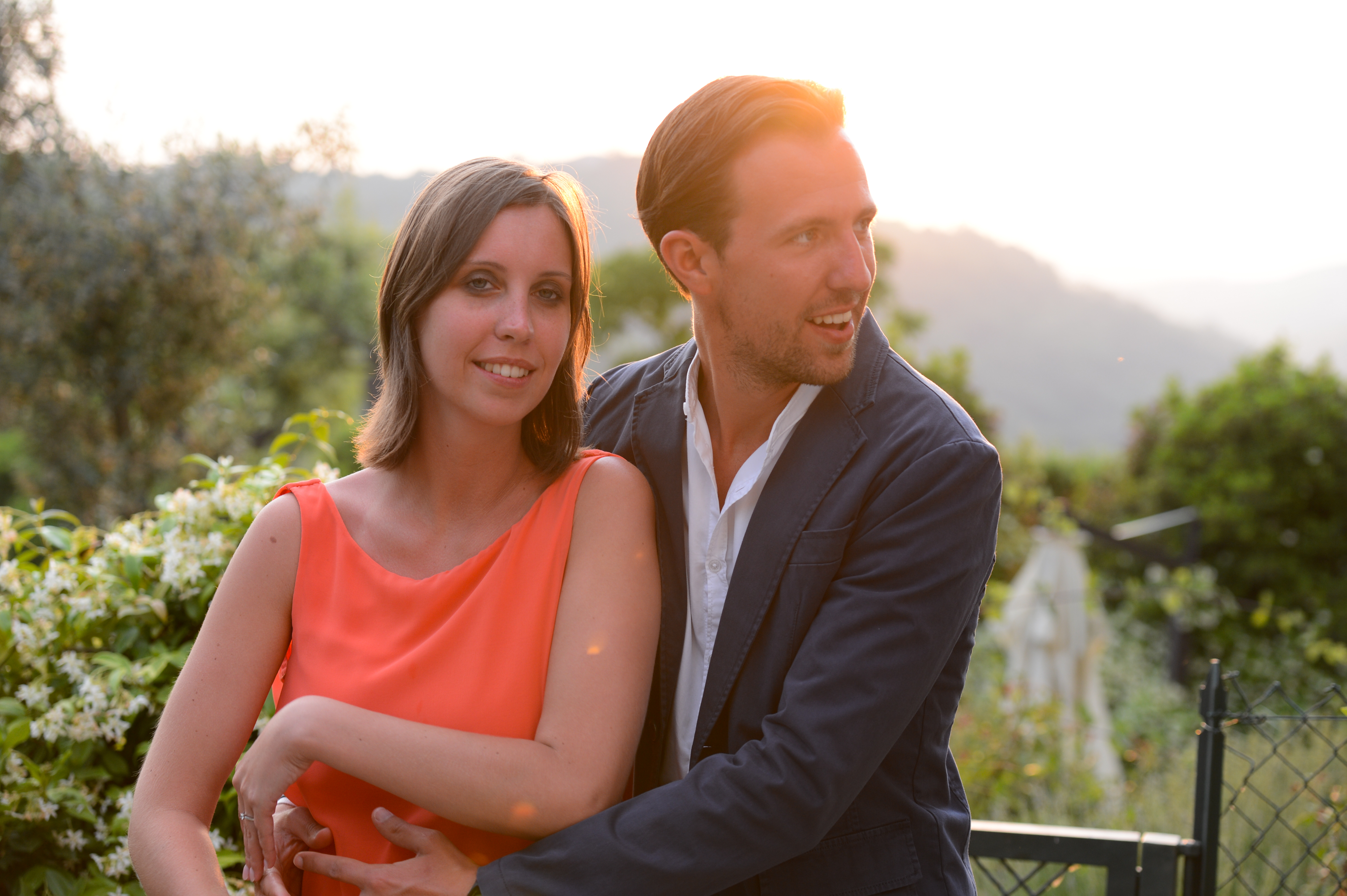 A couple poses together outdoors, with the man embracing the woman from behind.